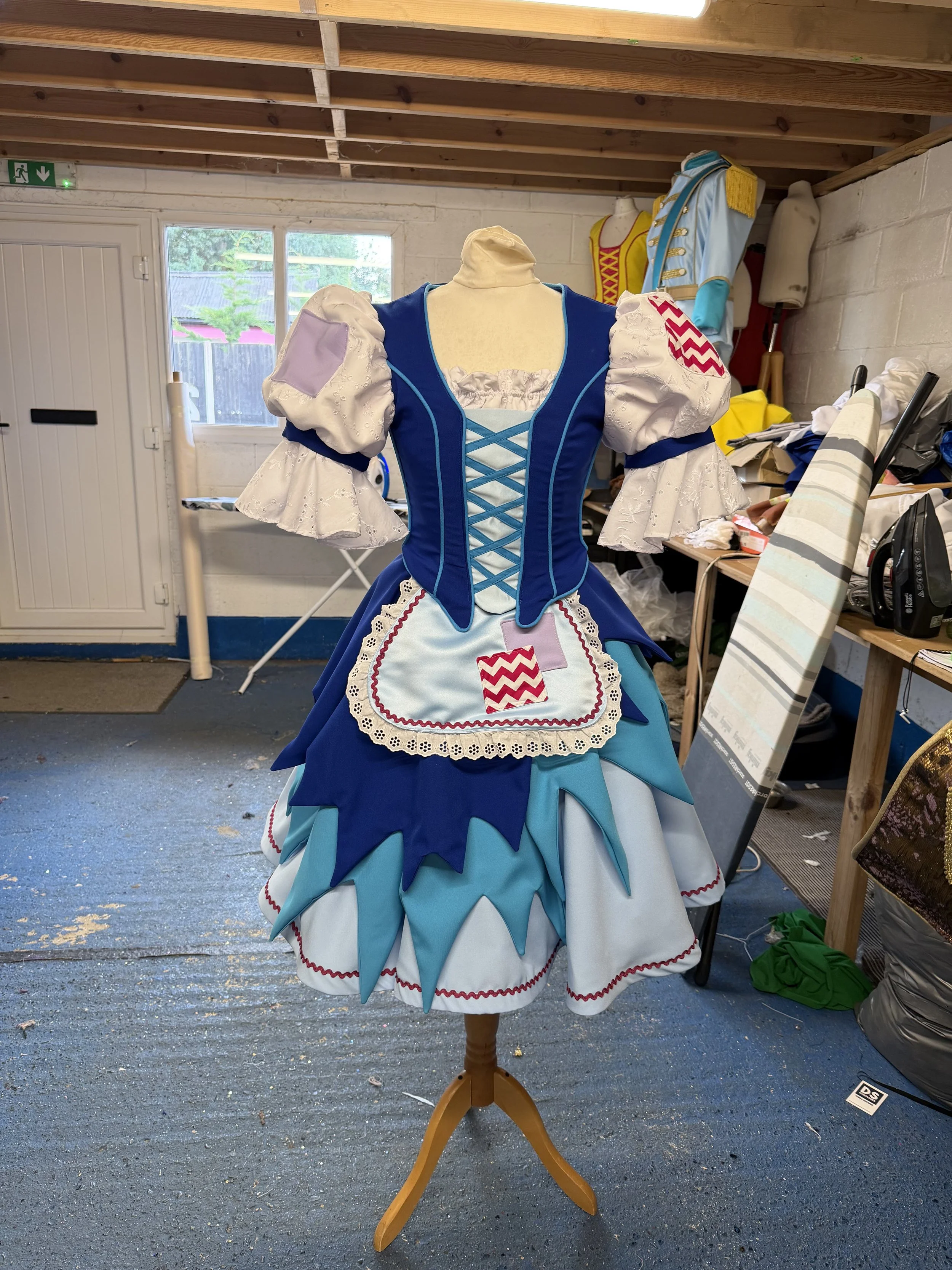 A colorful, elaborate costume dress displayed on a mannequin stand inside a workshop or storage room, with various fabric and clothing items in the background.