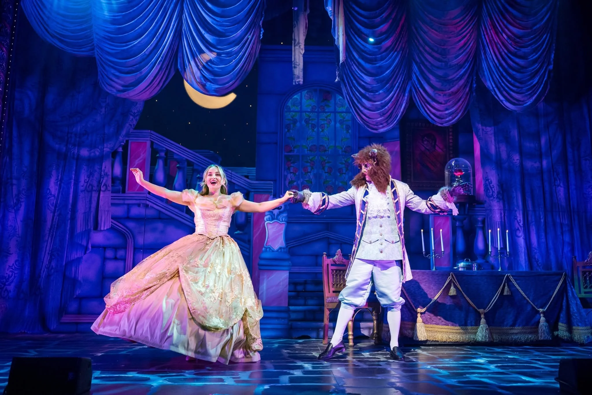 Stage scene from a theatrical production showing a woman in a pink and gold ball gown and a man in a princely costume holding hands and dancing against a backdrop of a blue curtain, moon, stars, and a window with floral designs.