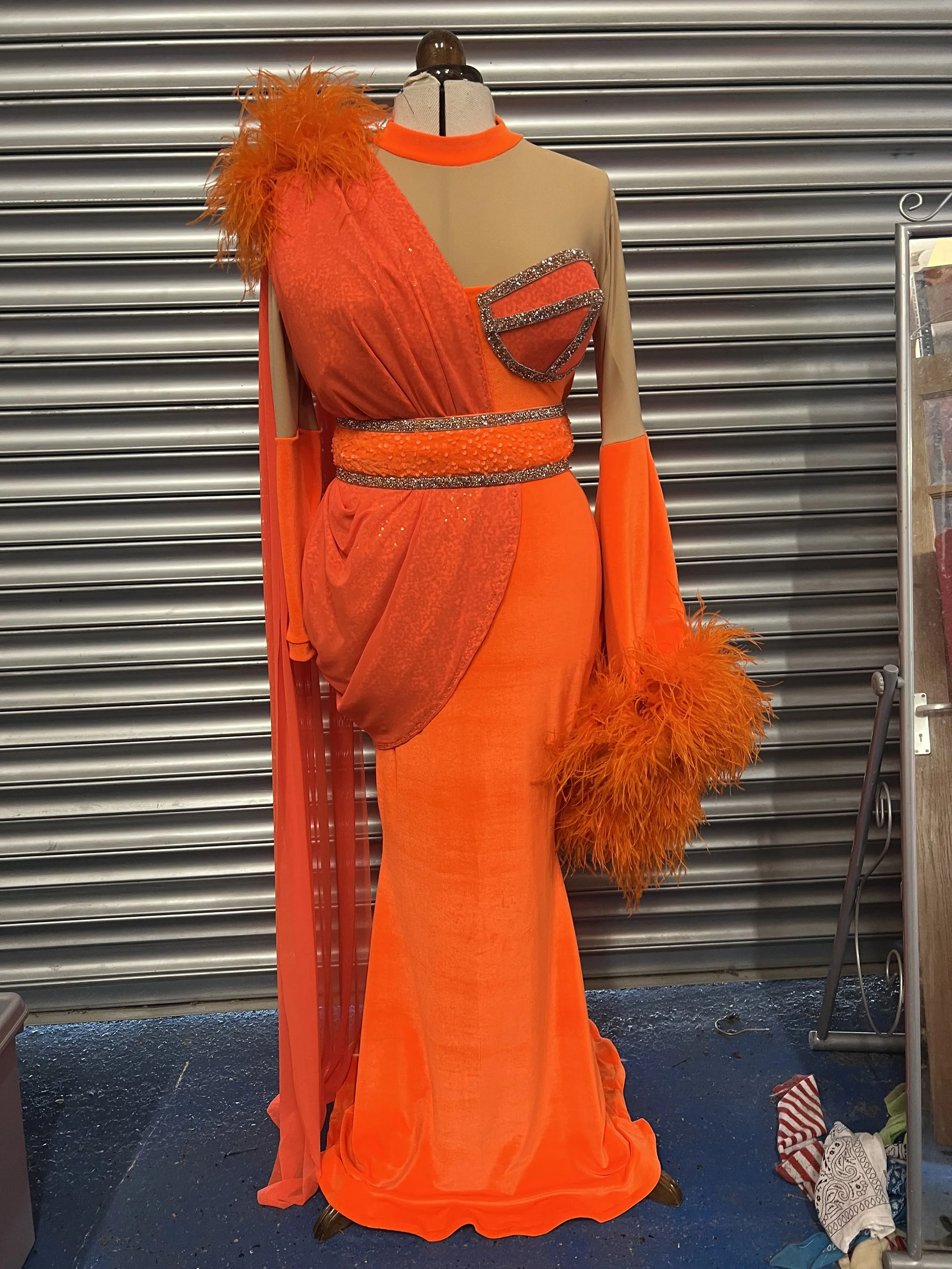 A mannequin displays a vibrant orange gown with sheer and textured fabric, embellished with rhinestones, and feathered accents on the shoulder. The gown features long sleeves and is accessorized with a matching orange feathered clutch.