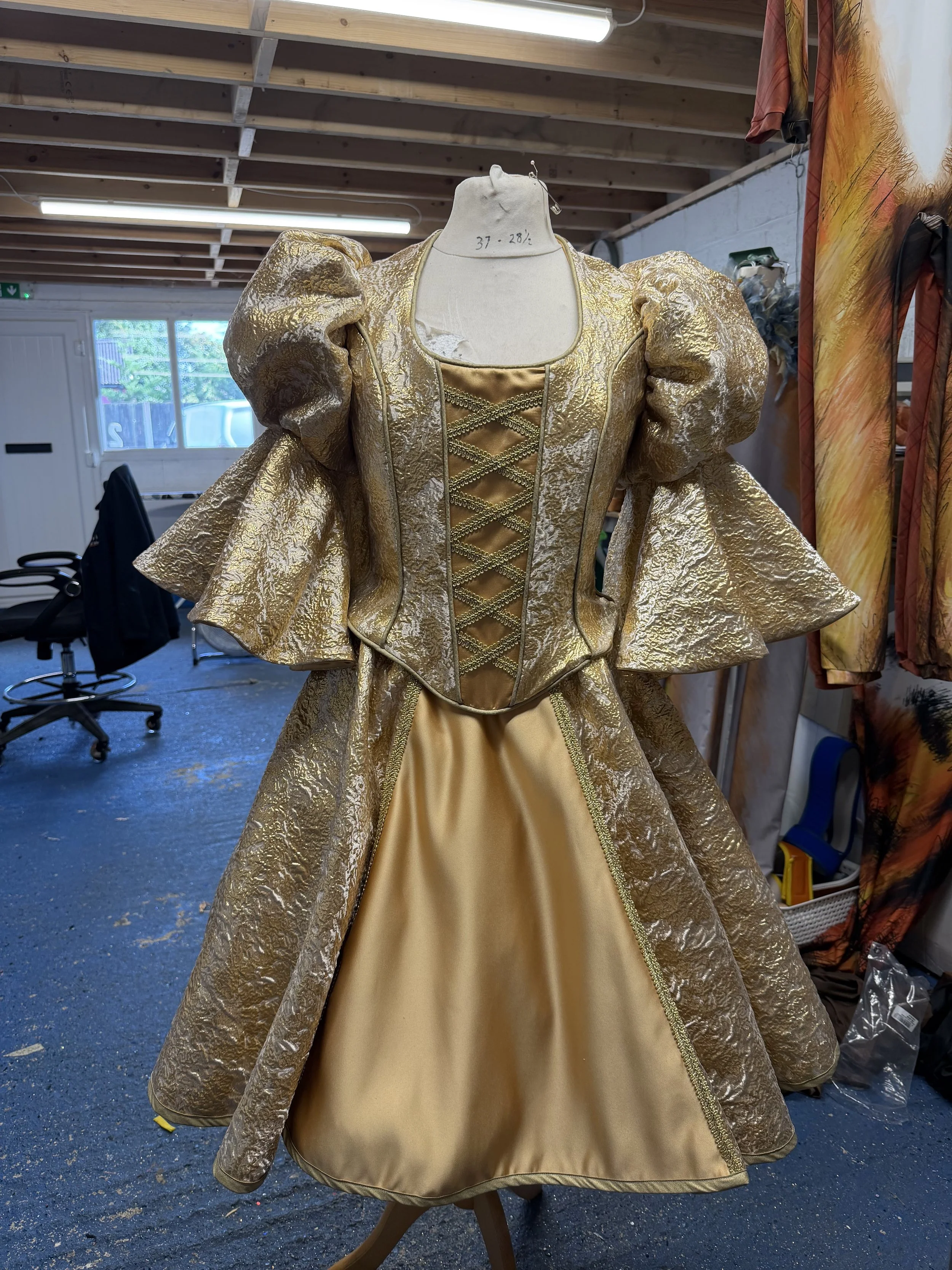 A gold and beige dress with puffed sleeves and intricate lace design, displayed on a mannequin in a workshop or costume room.
