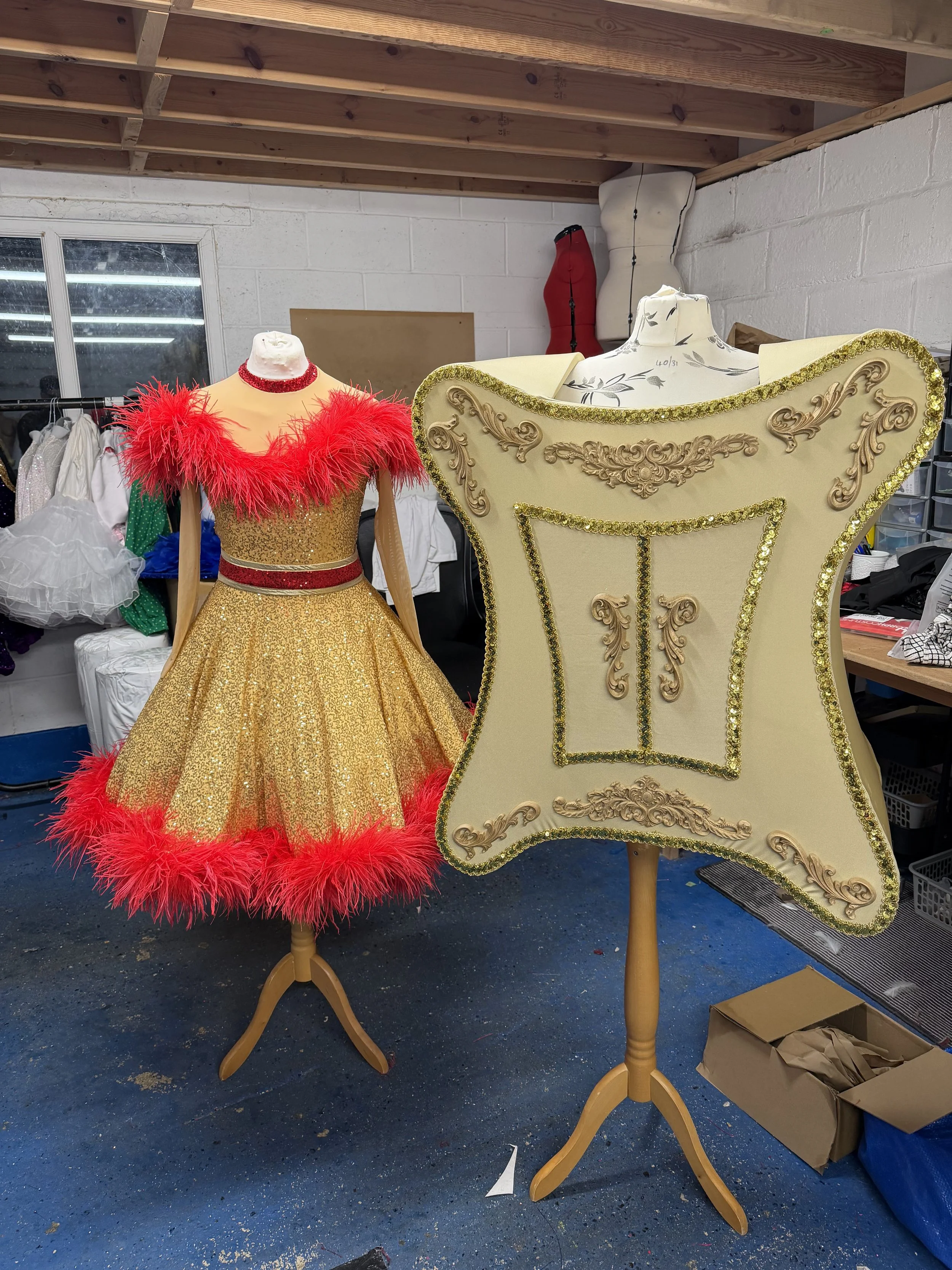 Two mannequins on stands dressed in costumes, one in a gold dress with red feather trim and the other in a large cream-colored dress with gold decoration, in a workshop or storage room with various clothing and materials around.