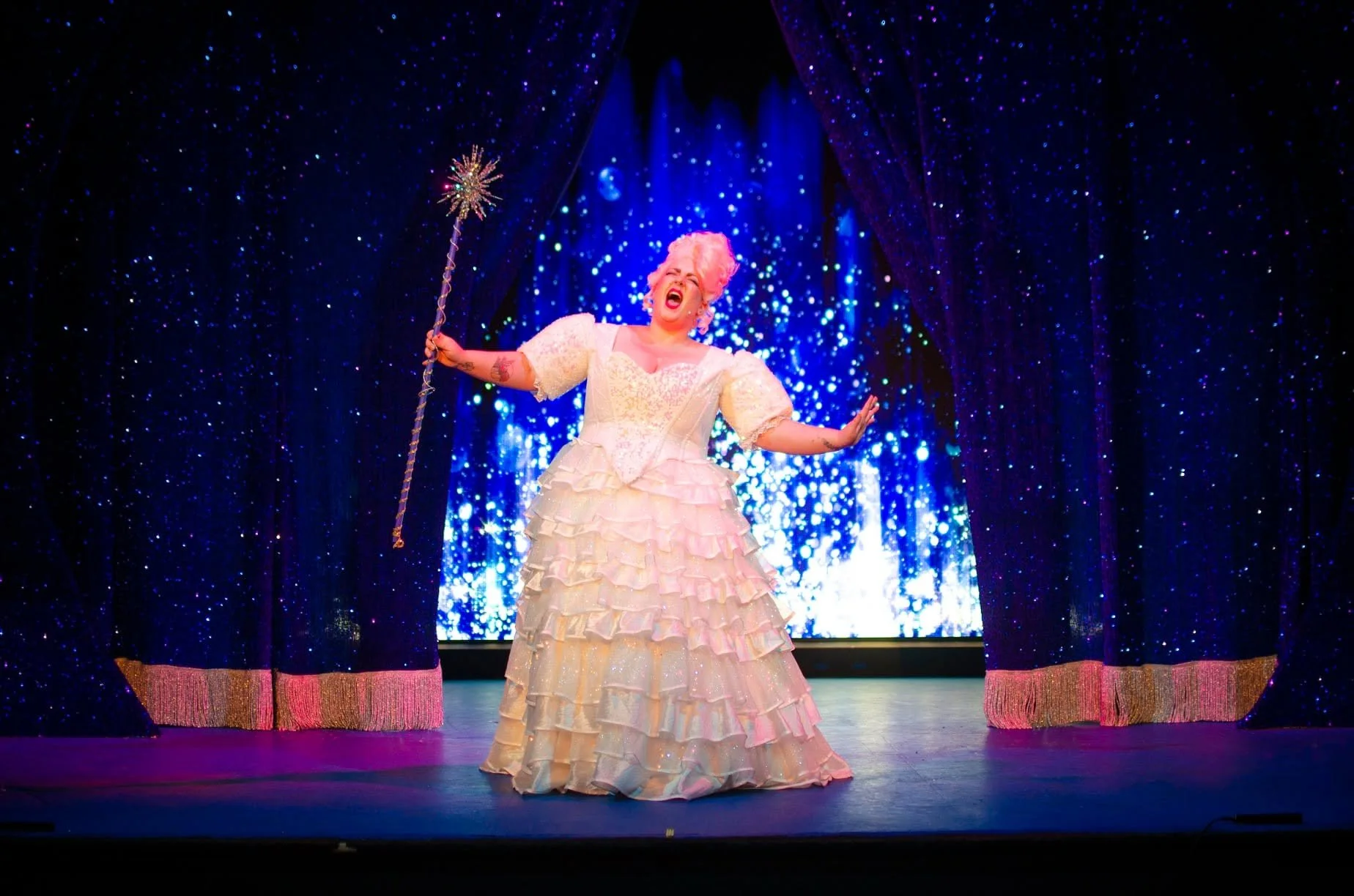 A performer dressed in a pink, ruffled, off-the-shoulder gown singing on stage, holding a sparkler, with a sparkling blue curtain backdrop.