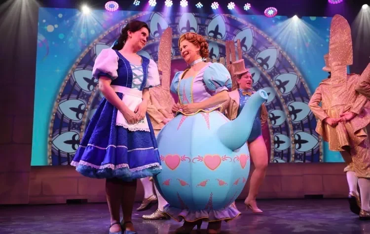 Stage production of Disney's Alice in Wonderland featuring actors in colorful costumes, including one dressed as a teapot, with a whimsical backdrop and stage lights.