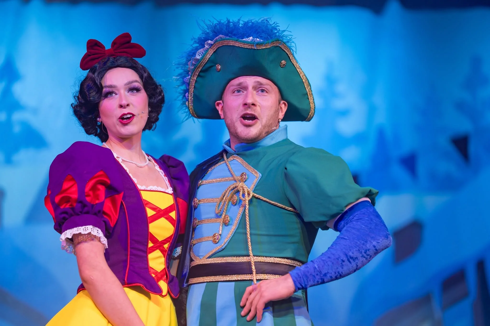Two performers in colorful costumes on stage. The female performer wears a purple and yellow dress with red accents and a large red bow in her hair; the male performer wears a green and blue uniform with gold trim and a large green hat with feathers. They appear to be singing or speaking, with a blue background behind them.