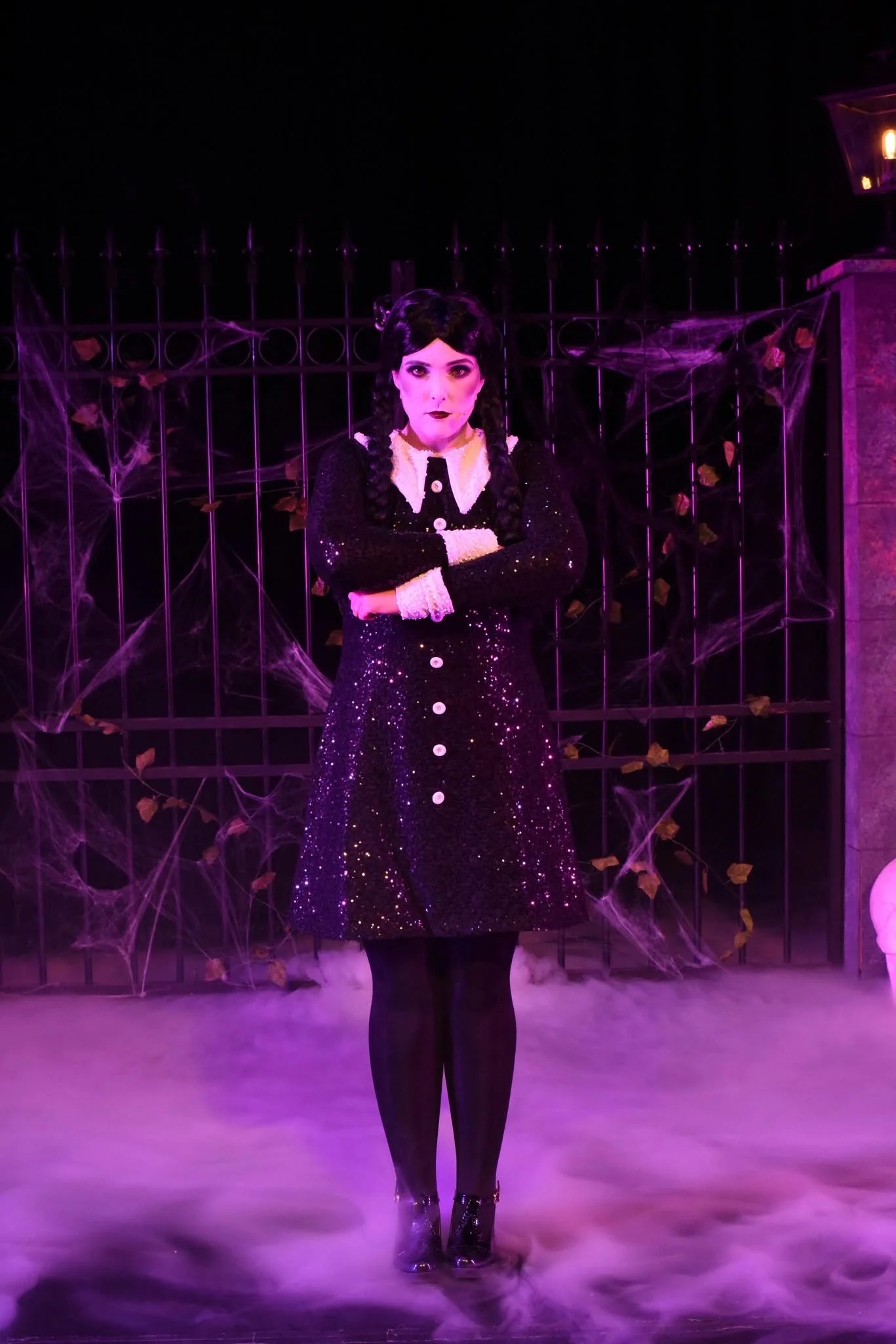Woman dressed in a sparkly black dress with white collar and buttons, standing with arms crossed in front of a spooky background with spider webs and fallen leaves, illuminated with purple lighting.
