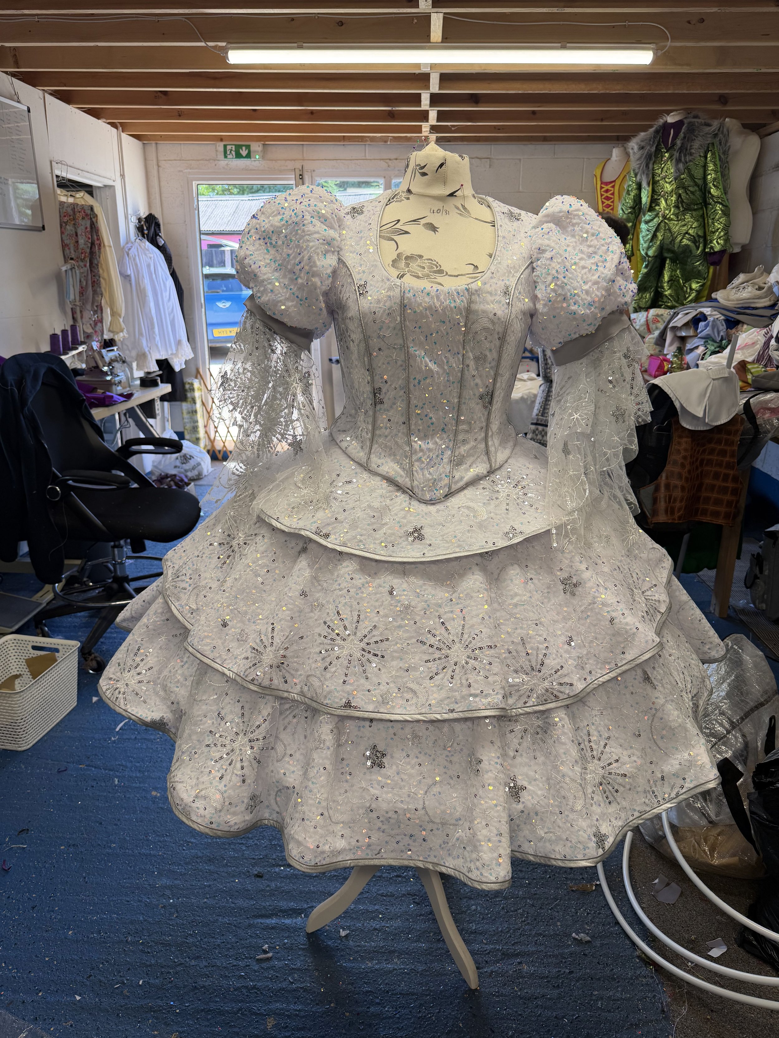 A white mannequin dressed in an elaborate, sparkling wedding gown with puffed sleeves, detailed embroidery, and a layered skirt, placed on a table in a cluttered backstage or costume shop with various clothing and fabric items around.