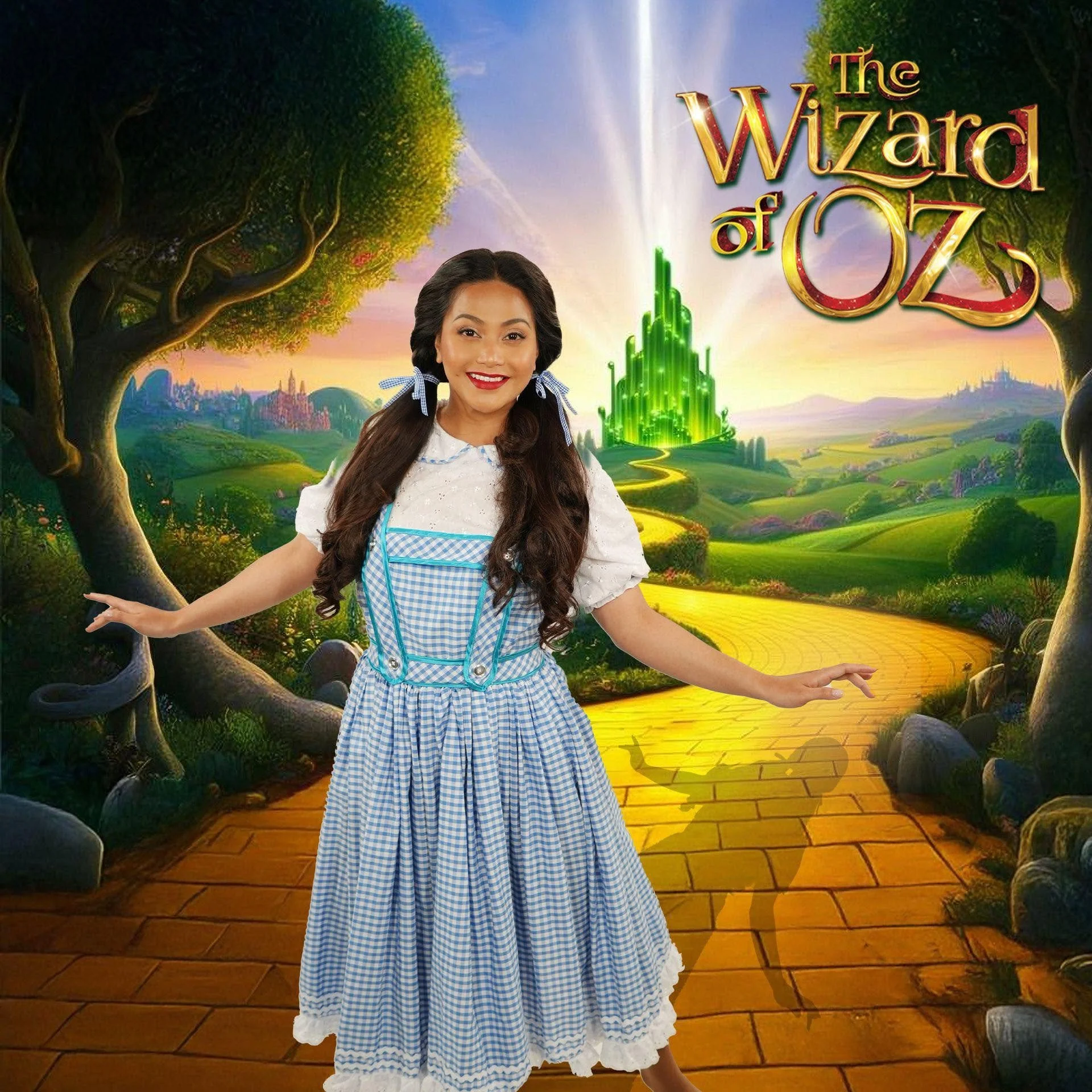 A young woman in a blue and white gingham dress with puffy sleeves and a white collar, smiling and standing with her arms outstretched in front of a colorful painted backdrop of a fantasy landscape. The backdrop features a yellow brick road, green tr