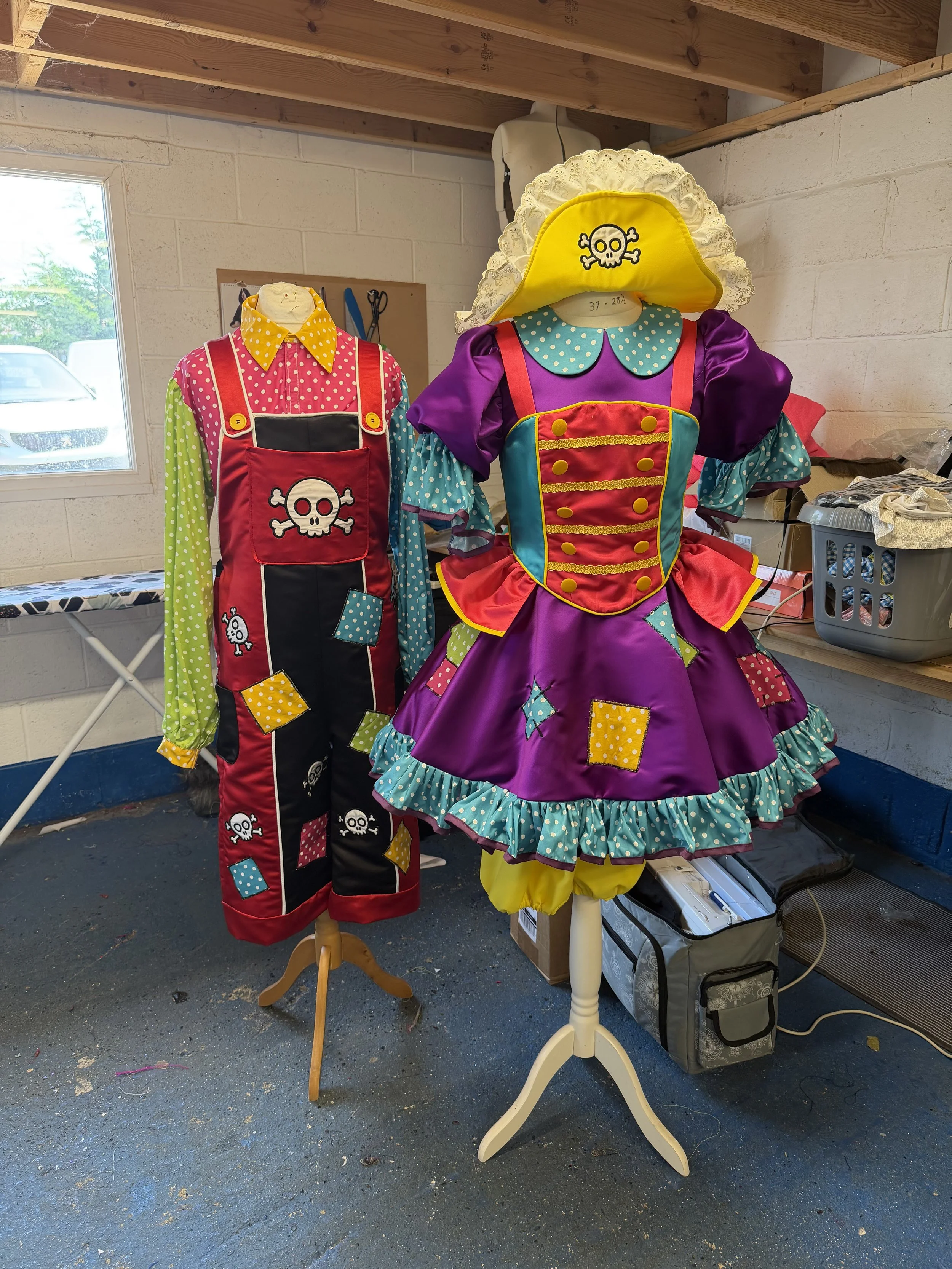 Costumes of two cartoon characters with skull and crossbones designs, one in simple clothing and the other in a colorful dress, displayed on mannequins in a room with a window, an ironing board, and storage boxes.