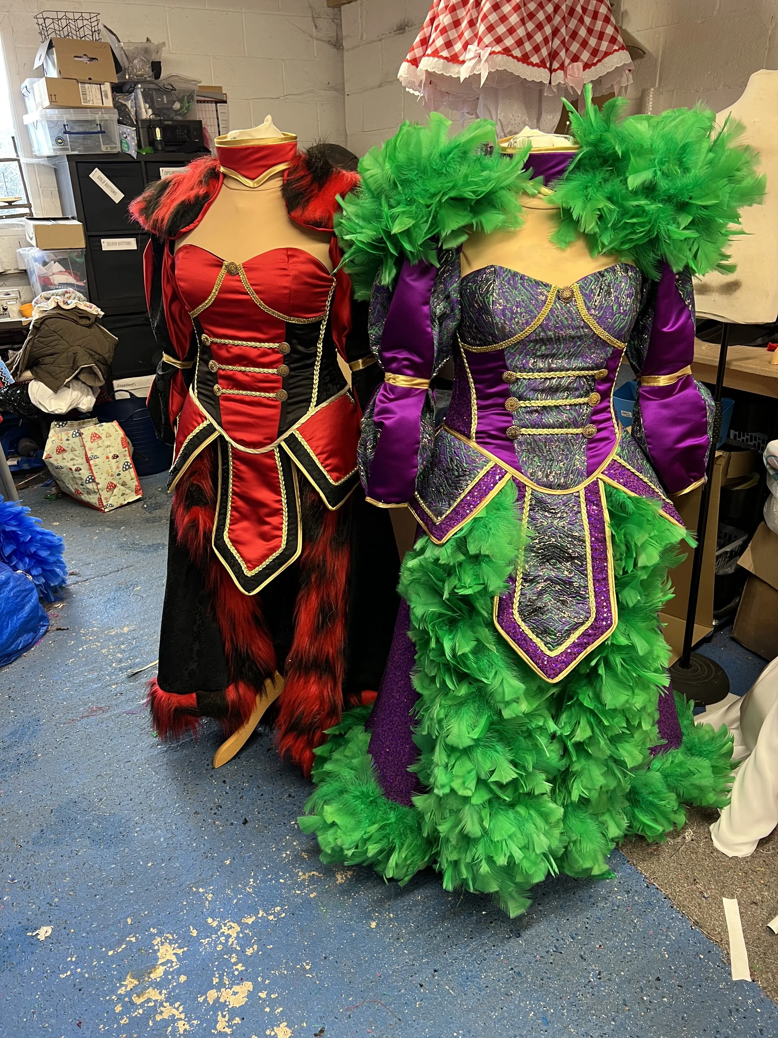 Two colorful costumes on mannequins in a cluttered room. One costume is red and black with gold accents, fur trim, and a cape. The other is purple and green with feathers, gold accents, and detailed fabric.