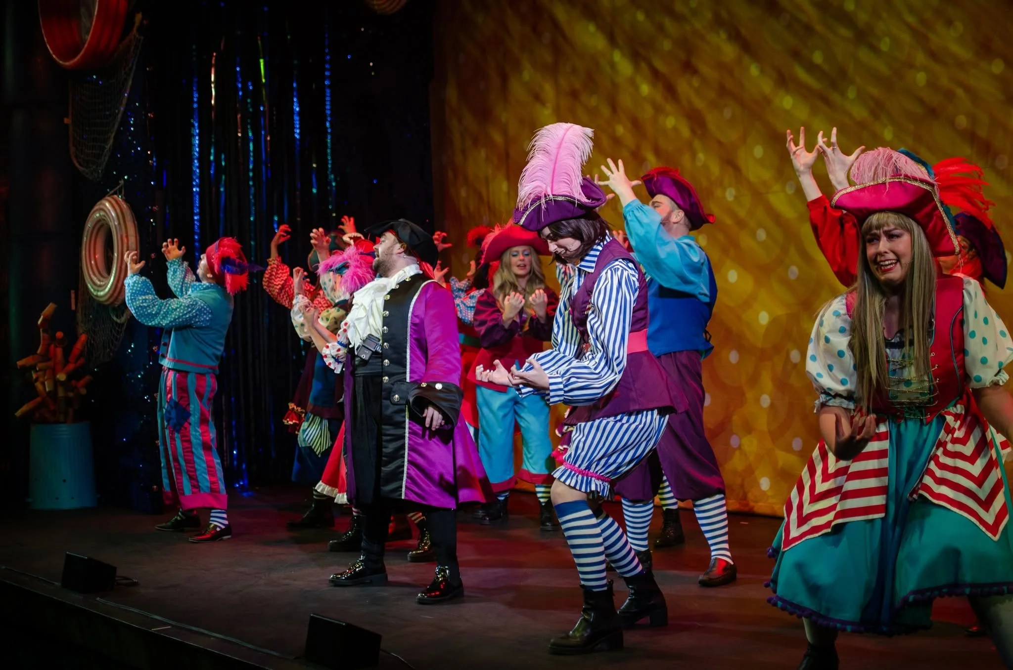People dressed in colorful, clown-like costumes with striped pants, feathered hats, and bright clothing are performing on a stage with a gold background and shiny decorations.
