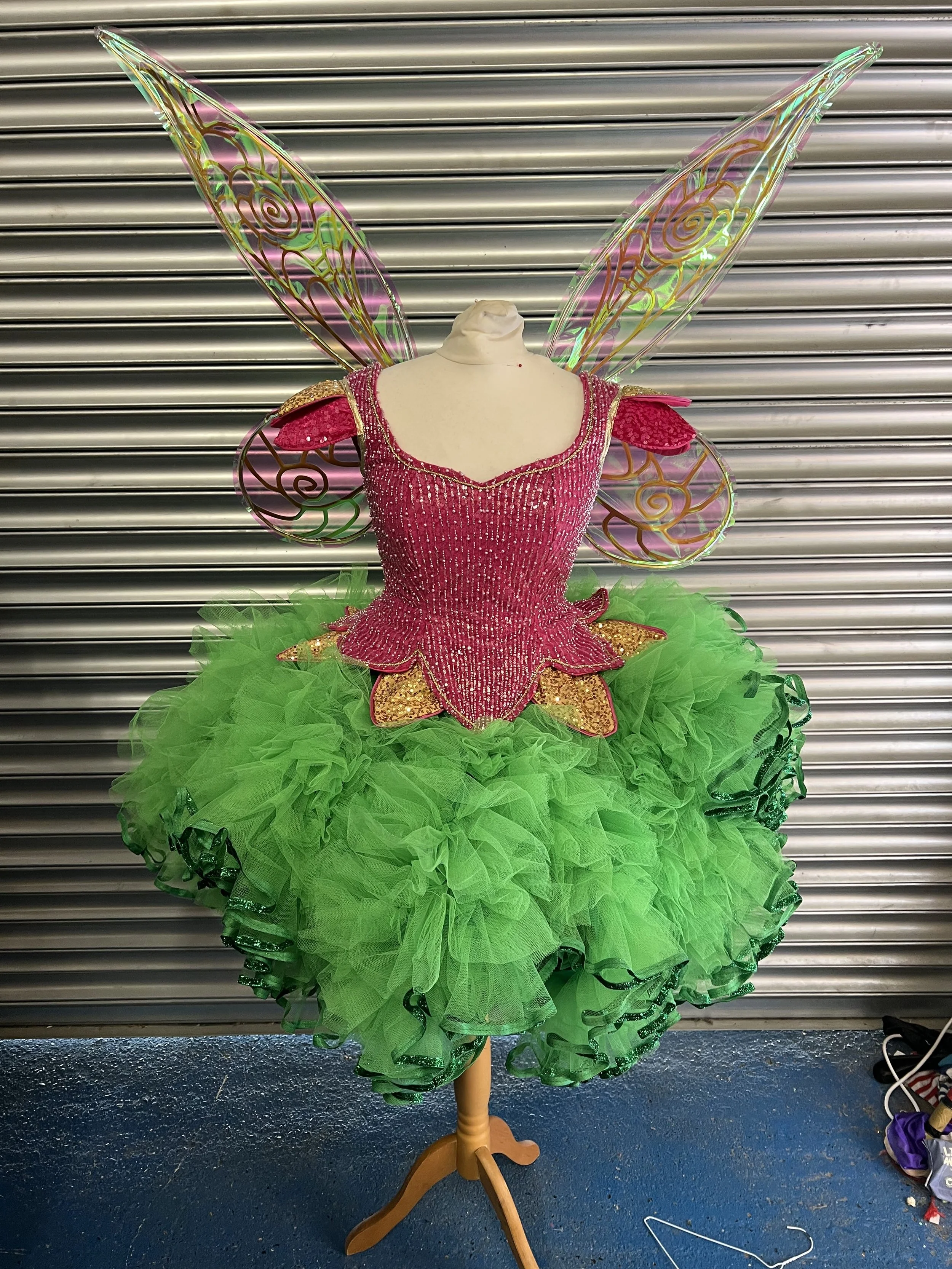 A costume on a mannequin resembling a fairy or butterfly with large iridescent wings, a pink bodice decorated with sequins, and a bright green tutu.