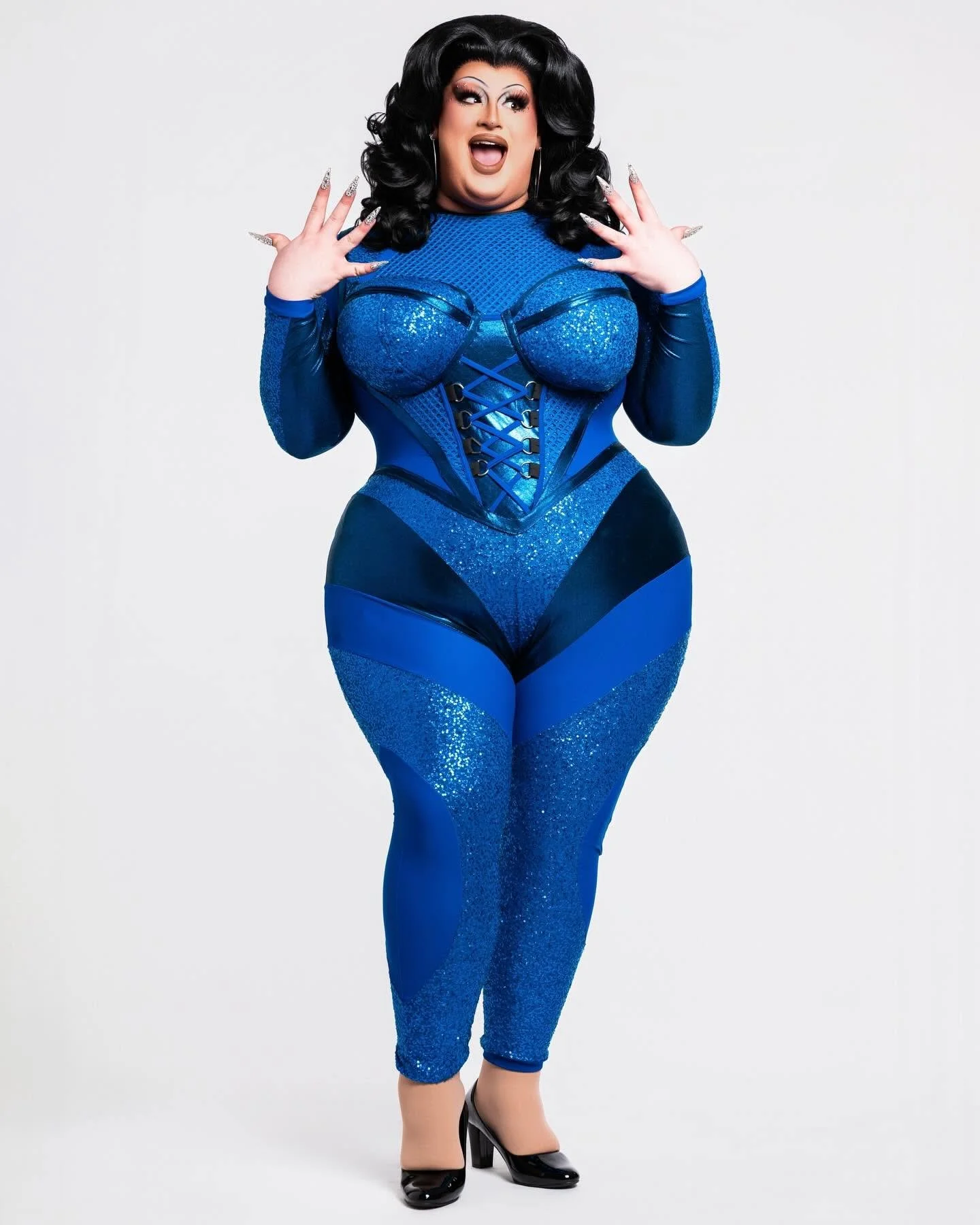 A plus-sized woman with dark hair styled in large curls, wearing a sparkly blue bodysuit with black accents, high heels, and statement makeup, posing in front of a plain gray background.