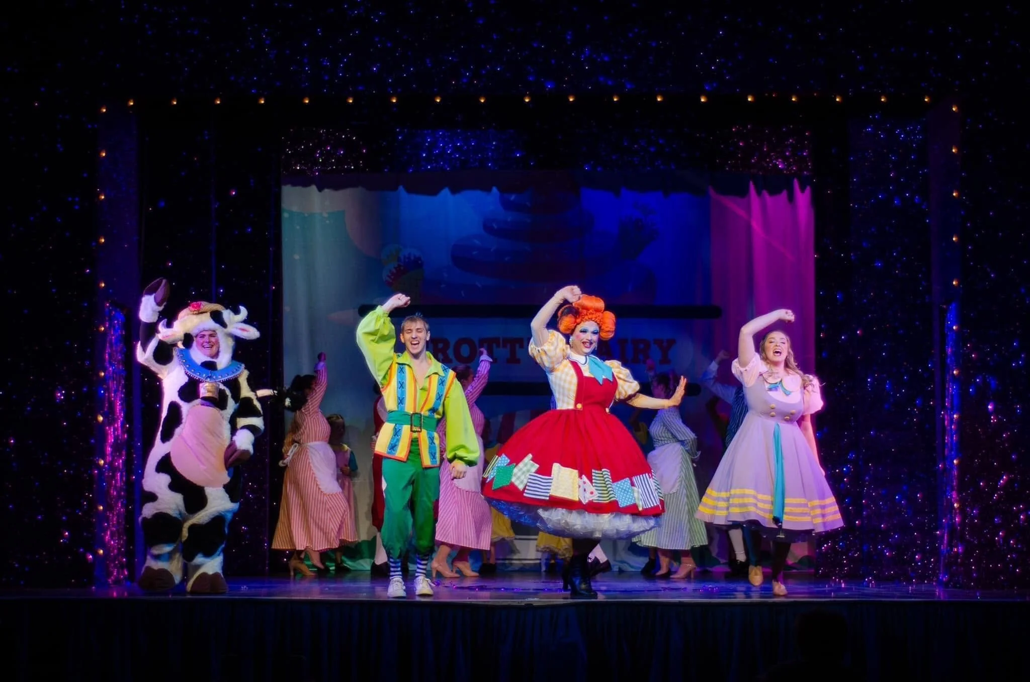 Stage performance featuring actors in colorful costumes, including a character in a cow costume, and a woman in a red dress with jump ropes. The background has a starry theme with colorful curtains.