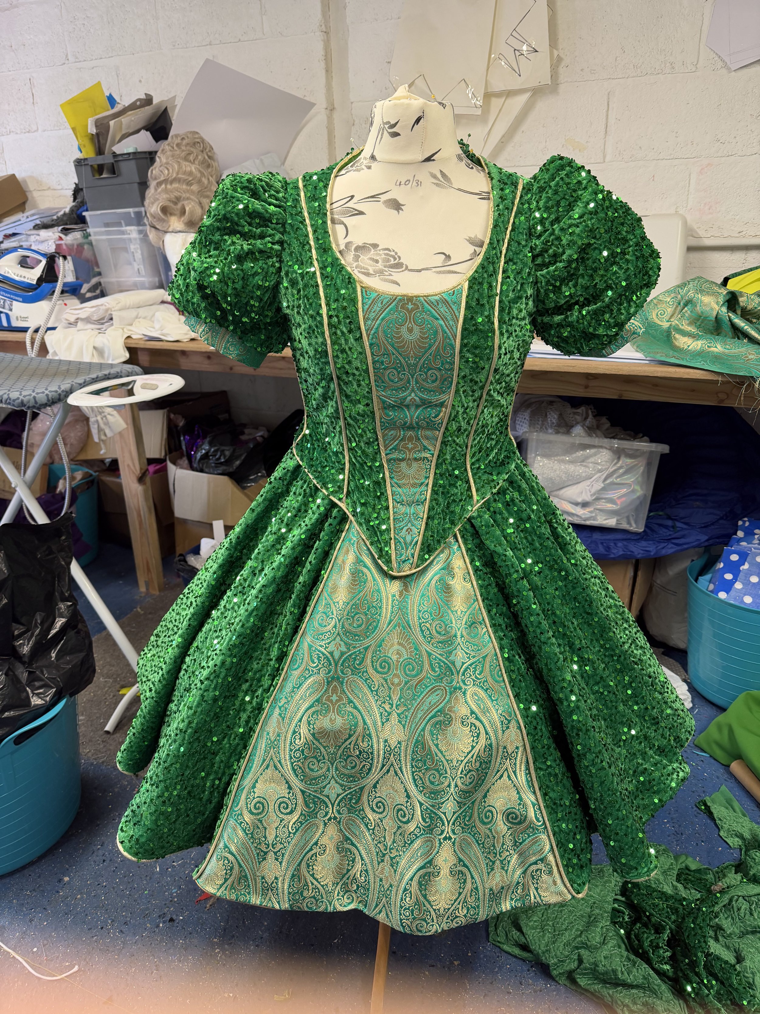 A green, sequined dress on a mannequin, with ornate gold and green fabric at the front and puffed sleeves, displayed in a cluttered workspace.