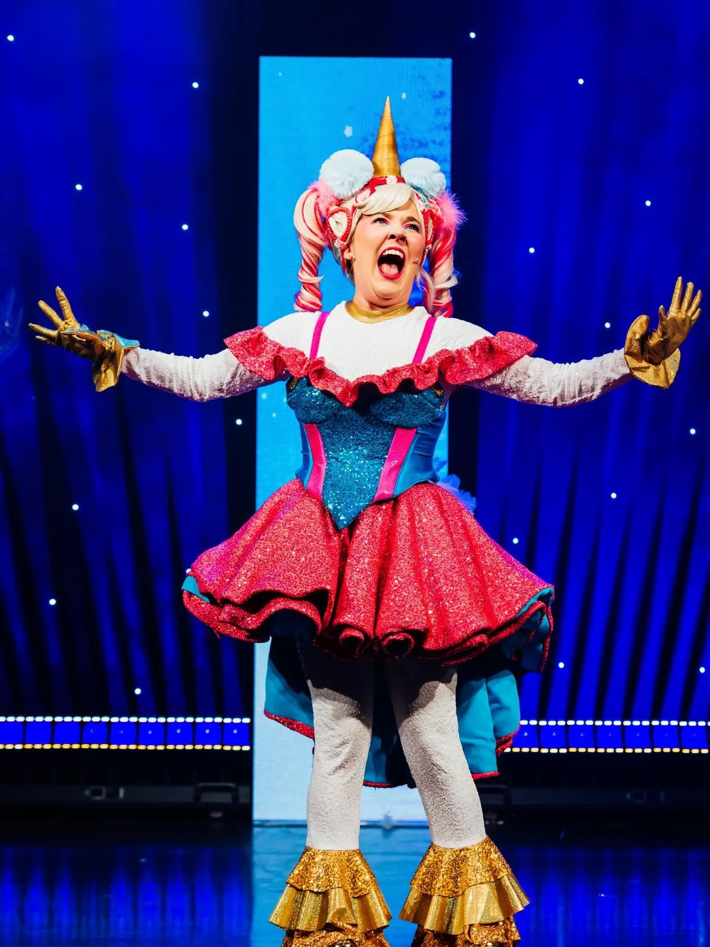 Performer dressed as a unicorn princess with a golden horn, pink and white hair, and colorful costume, singing on stage with a blue curtain and snowflake-like lights in the background.
