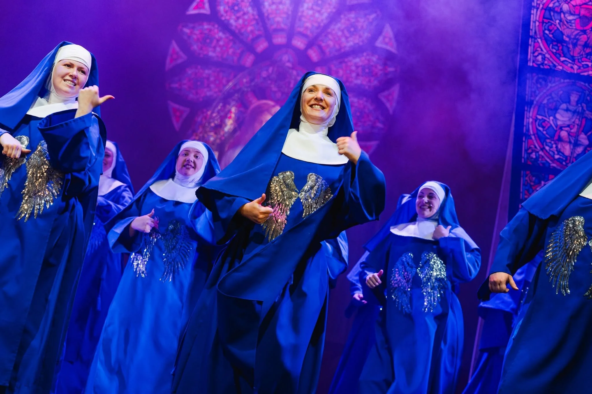 A group of women dressed as nuns performing on stage, smiling and dancing. They are wearing blue robes with silver eagle designs and white head coverings, with a large, colorful, stained-glass window backdrop.