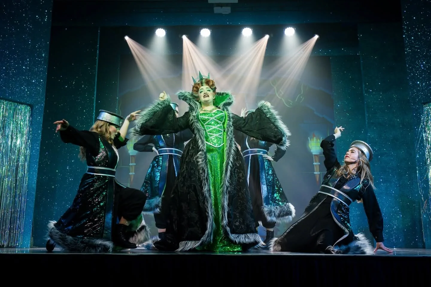 A theatrical stage performance featuring a central woman dressed as the Wicked Witch of the West from the Wizard of Oz, with bright lighting and backup dancers in black and blue costumes.