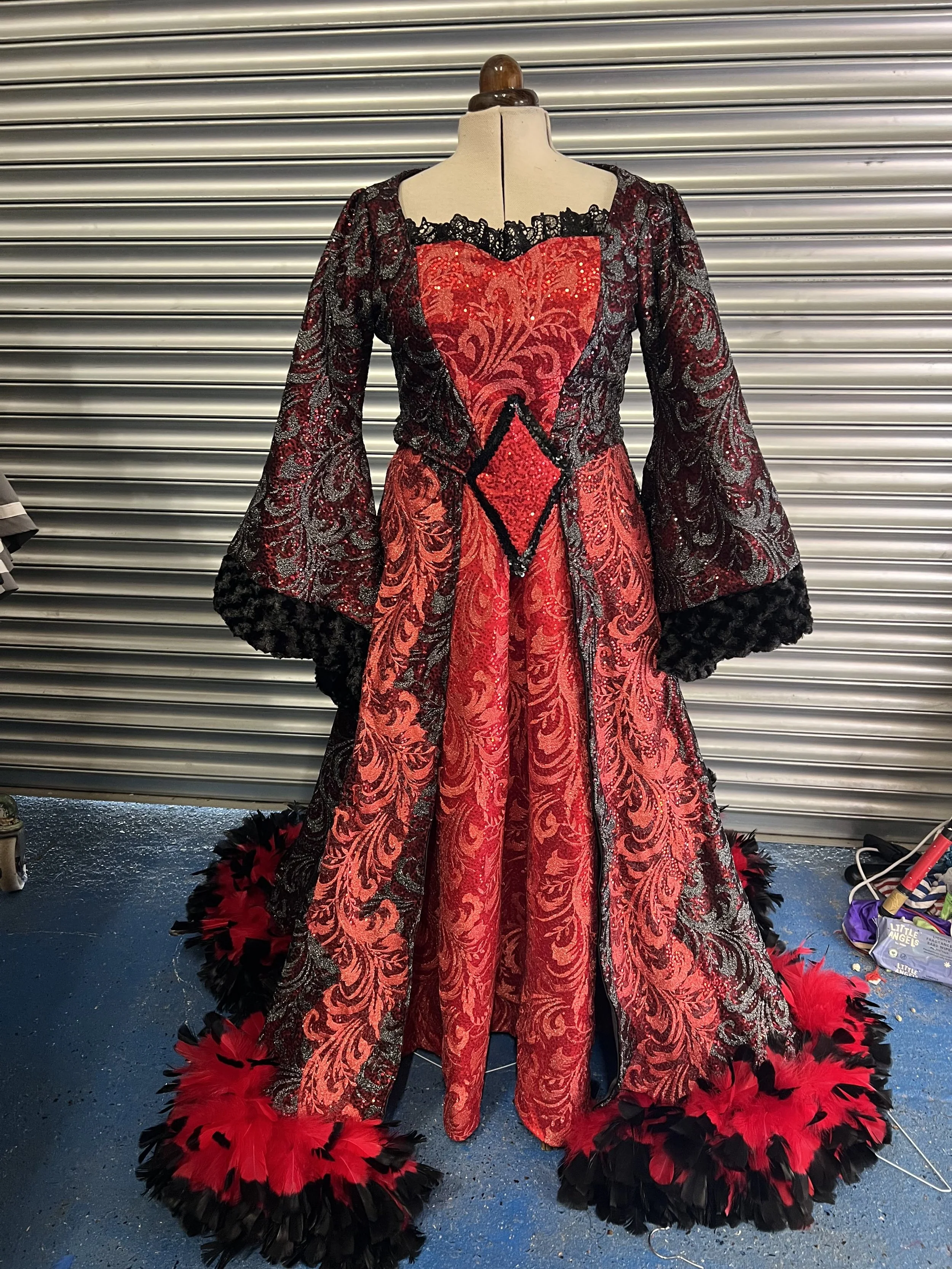 A costume dress featuring black and red lace fabric with intricate patterns, a heart-shaped bodice with gold sequins, and feathered trim at the hem, displayed on a mannequin against a metallic shutter background.