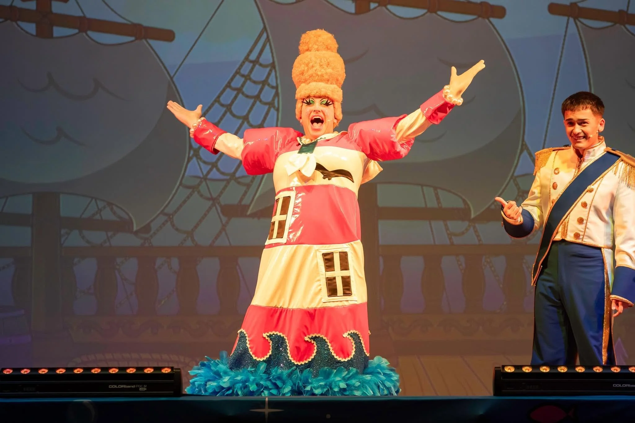 Performer dressed as a clown with arms outstretched and mouth open, standing on stage with a woman in a military costume beside her, in front of a colorful theatrical backdrop.