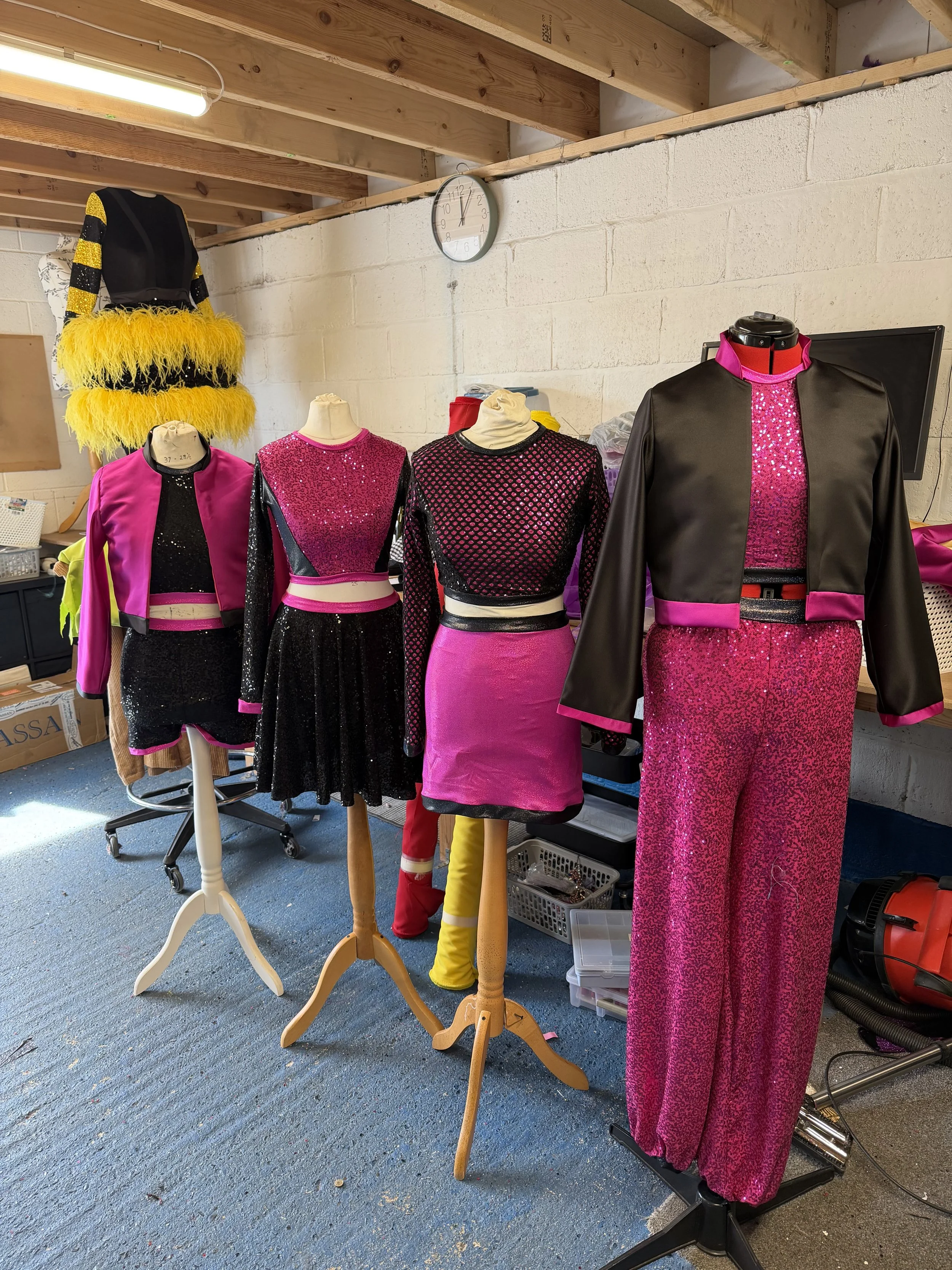 Mannequins displaying colorful costumes inside a room with a clock on the wall and exposed wooden ceiling beams.