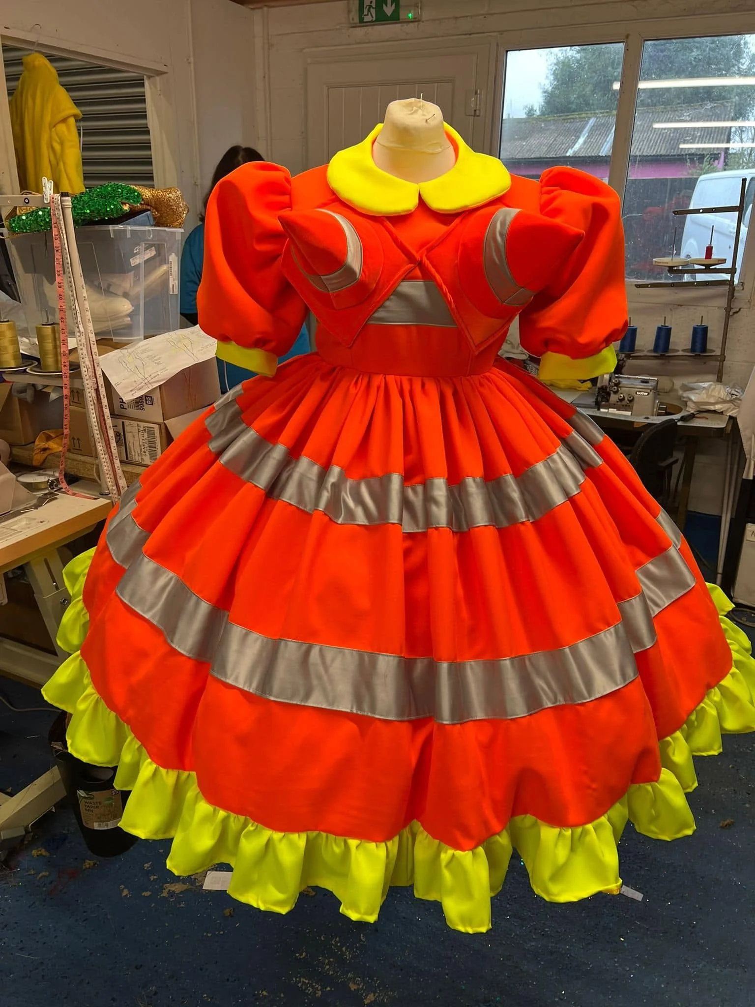 A colorful dress on a mannequin in a workshop. The dress is bright orange with yellow accents and silver reflective bands, with a large yellow collar and puffed sleeves. The skirt is wide and layered, with yellow ruffles at the bottom. In the backgro