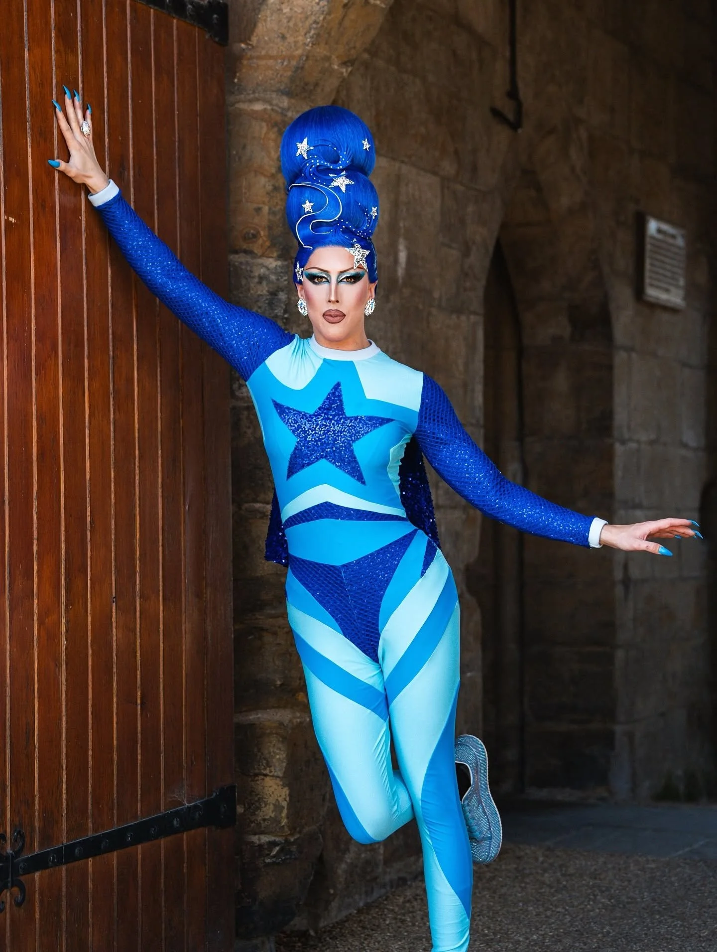 Person in a blue and white outfit with a star design, wearing elaborate blue hairstyle with star and moon decorations, posing against a wooden and stone background.
