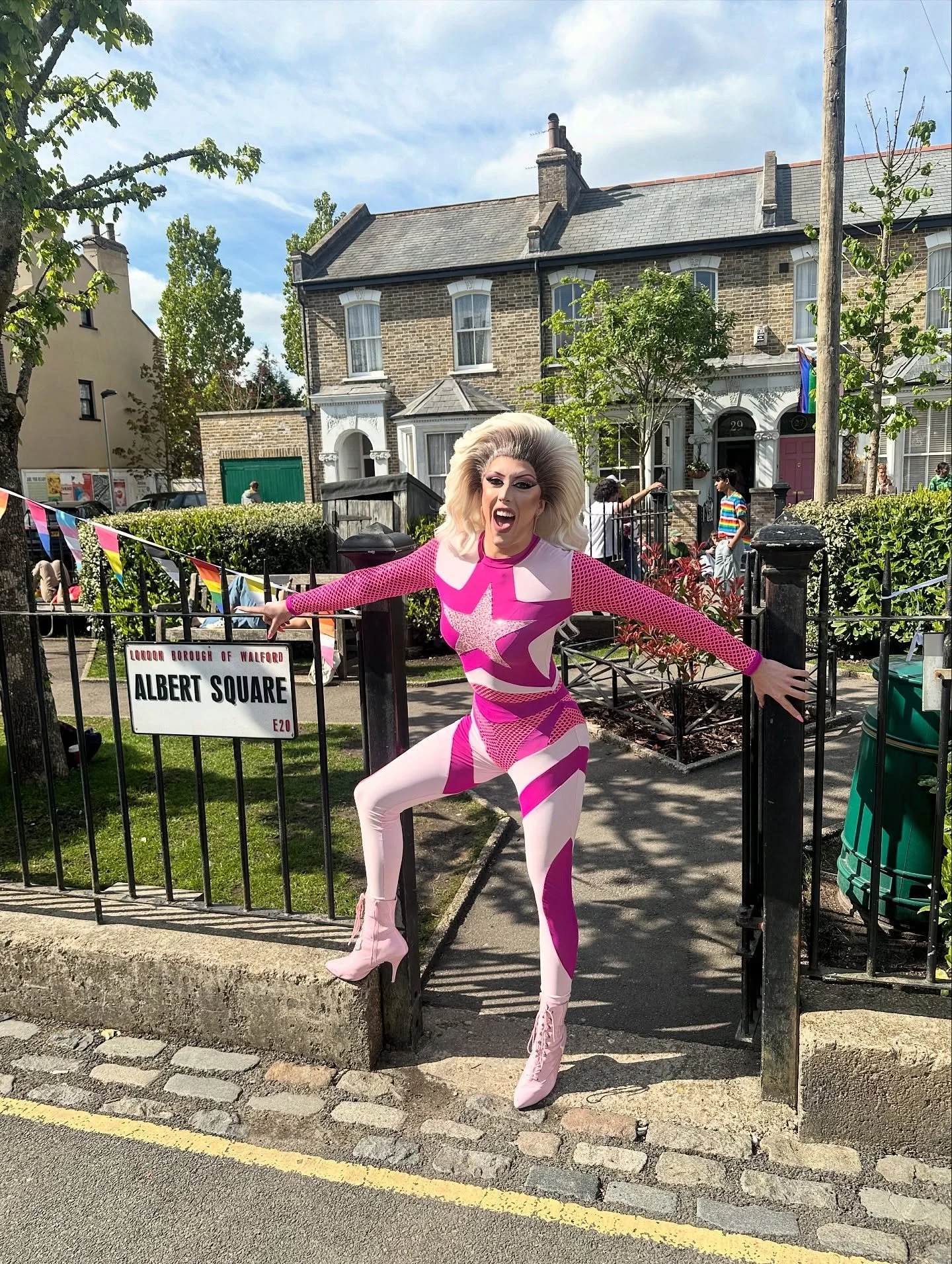 A person in a pink and white leotard with a star design, crimped platinum blonde hair, and pink heeled boots, standing behind a black fence at Albert Square in Walford, with Victorian houses and a few people in the background