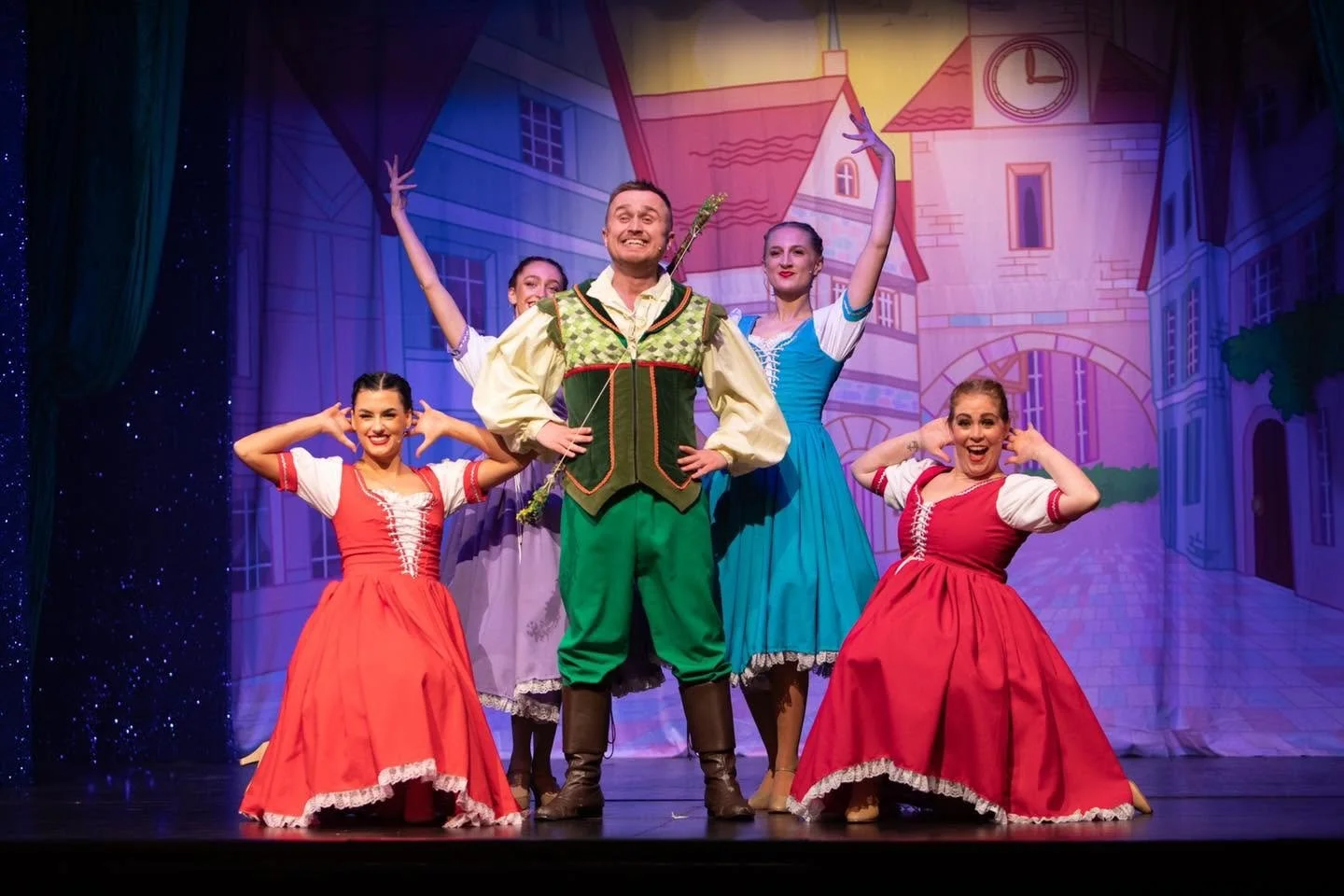 Theater performers dressed in colorful costumes on stage, with a backdrop of a cityscape and large curtains on sides. The central male performer is in green and beige, smiling confidently, flanked by four female dancers in red and blue dresses striking dance poses.