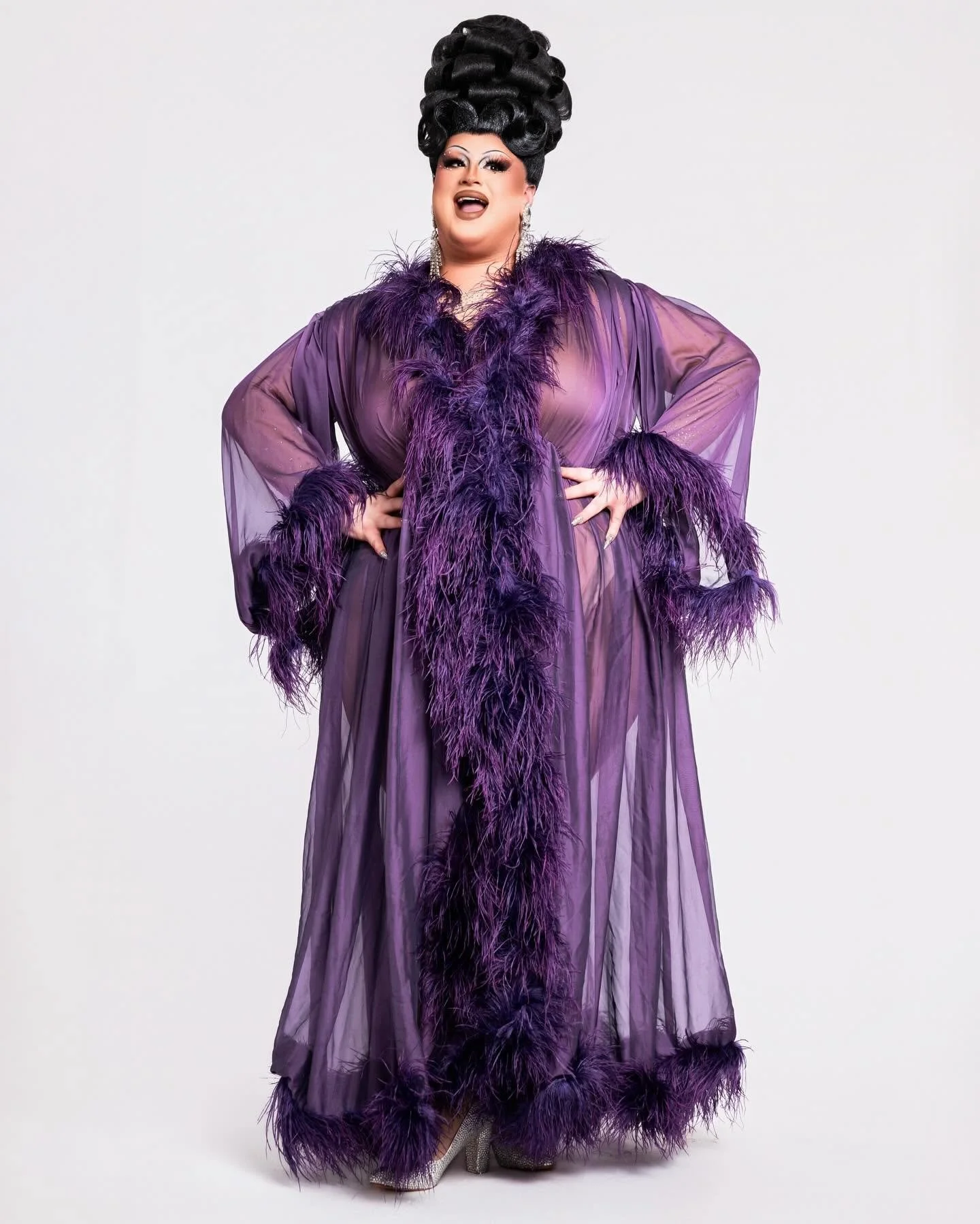 A person dressed in a long, sheer purple gown with purple feather trim, standing against a plain white background.