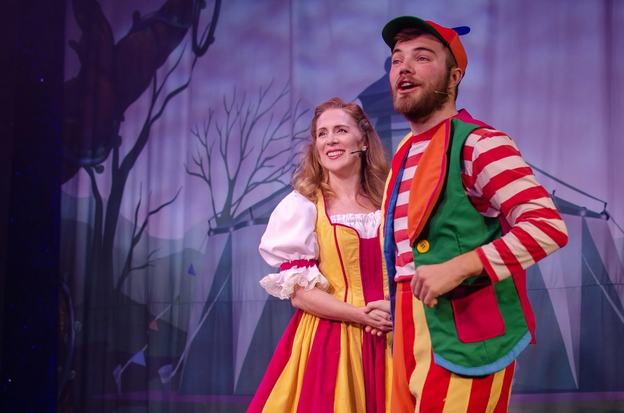 Two clowns, one woman and one man, stand on stage during a performance. The woman is smiling and wearing a white and yellow costume with a red and yellow apron, while the man is in a colorful, striped clown outfit with a green, red, and yellow vest and a red cap. The background features a purple curtain with a silhouette of trees.