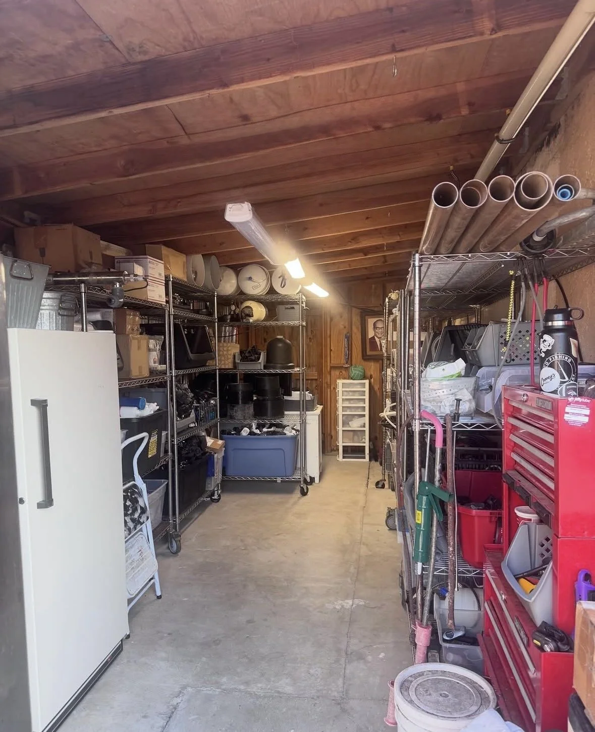 A well-organized garage with metal shelving units filled with various tools, storage bins, and household items. Pipes are stored on the top right shelf, and there is a framed picture on the wooden-paneled wall at the back along with a small drawer unit.