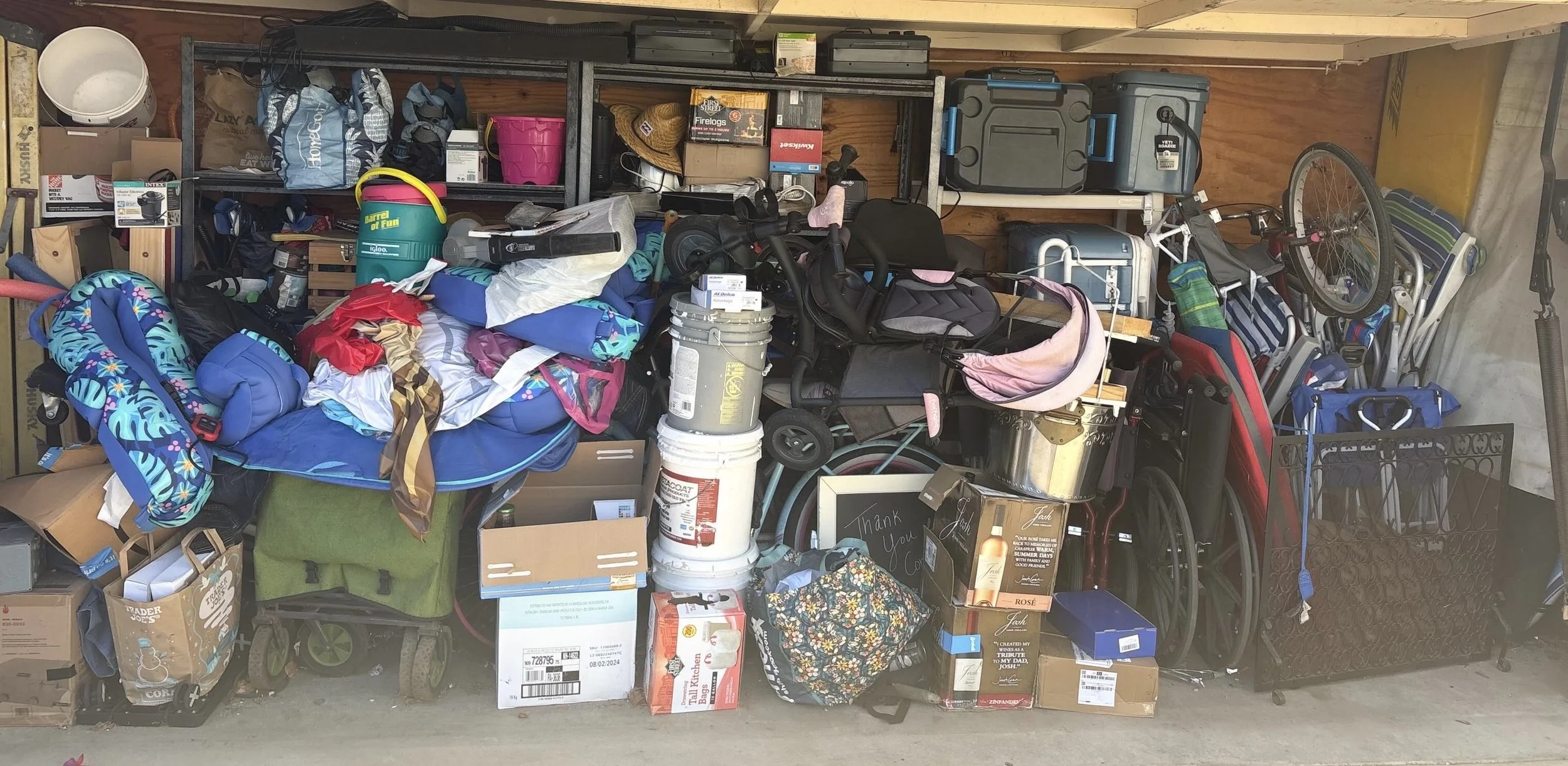 A cluttered garage filled with various items including a stroller, a wheelchair, boxes, bags, buckets, and gardening tools, with shelves holding containers, boxes, and a hat.