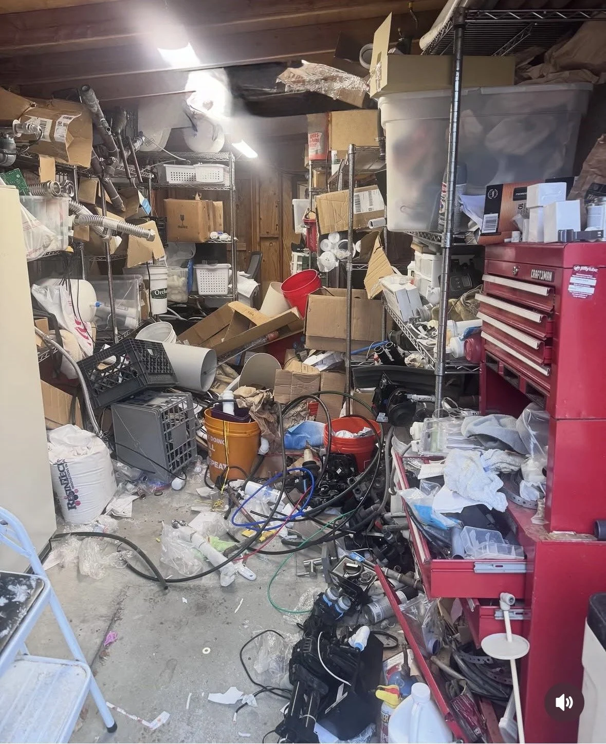 Cluttered garage with various tools, plastic bins, cardboard boxes, wires, and miscellaneous items scattered on the floor and shelves.