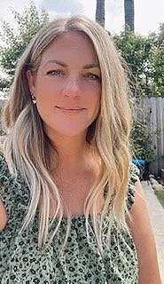 A woman with long, wavy blonde hair, wearing a patterned top, standing outdoors with trees and a fence in the background.