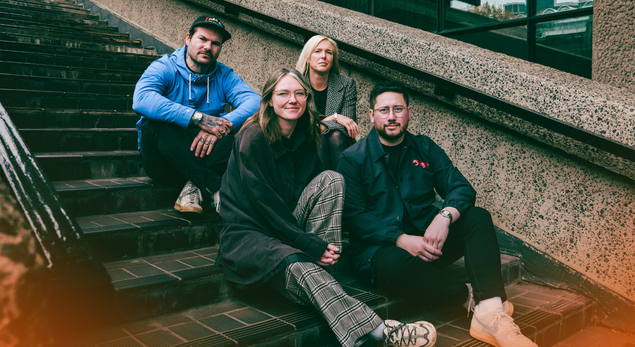 Four people sitting on black outdoor stairs next to a textured concrete wall, posing for a photo. Two women and two men, casually dressed, with a cityscape reflected in the glass window behind them.