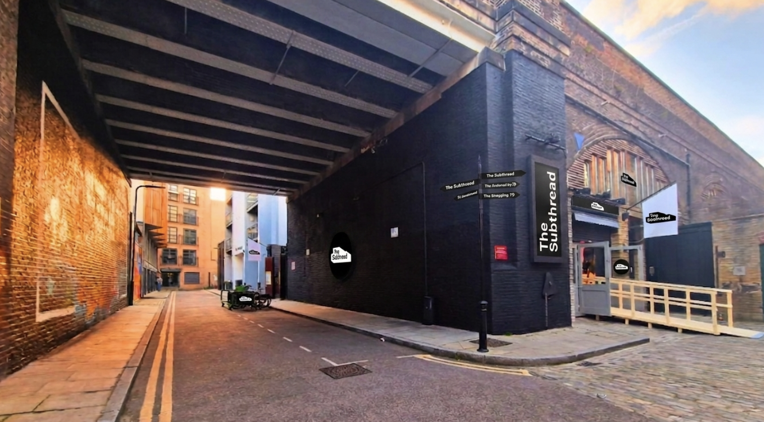 An urban alleyway with brick walls, a dark building under a railway, and a small ramp leading to a door. This is the venue for Inspire
