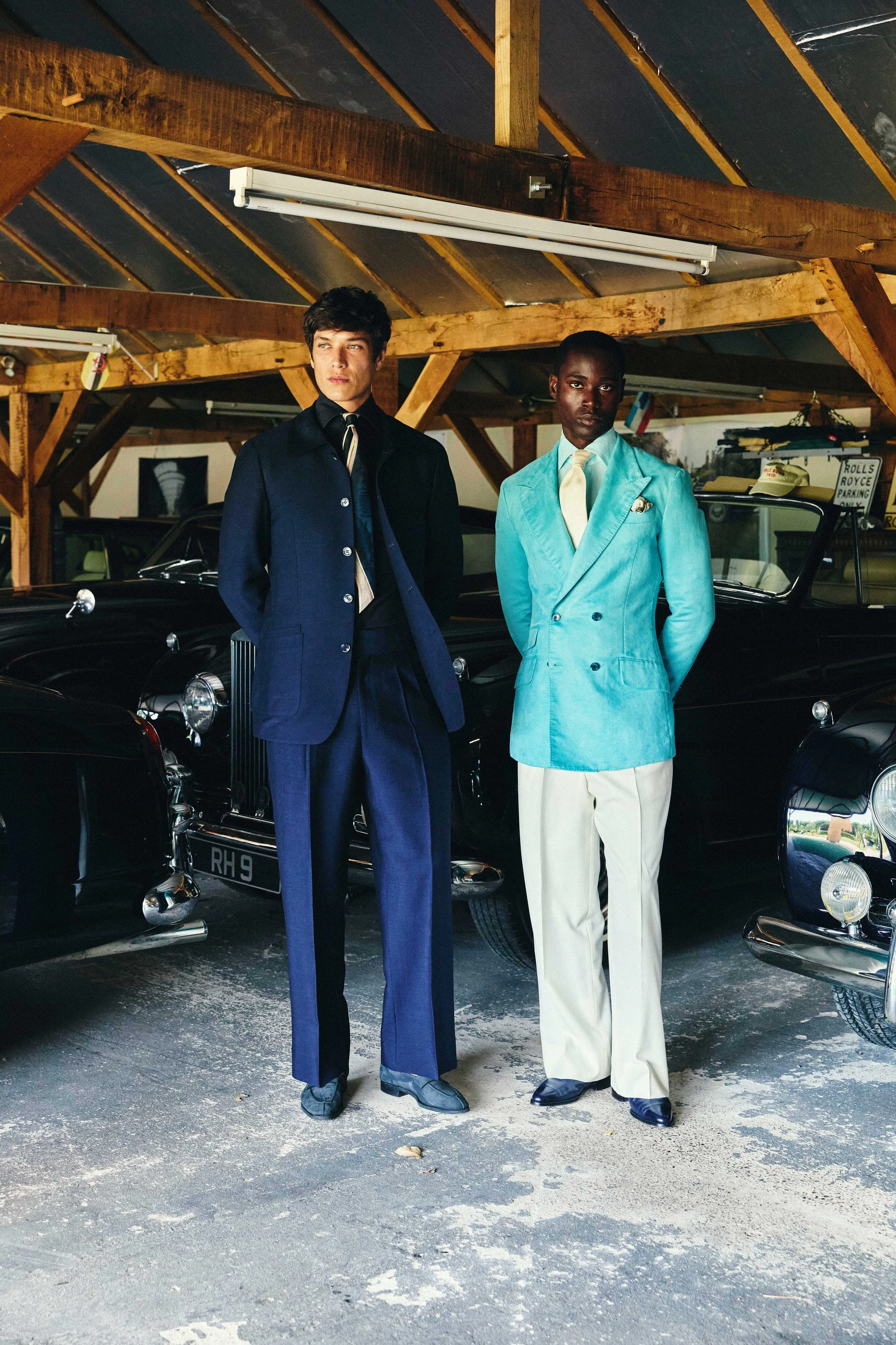Two men standing in a garage in front of vintage cars, dressed in formal suits and looking serious.