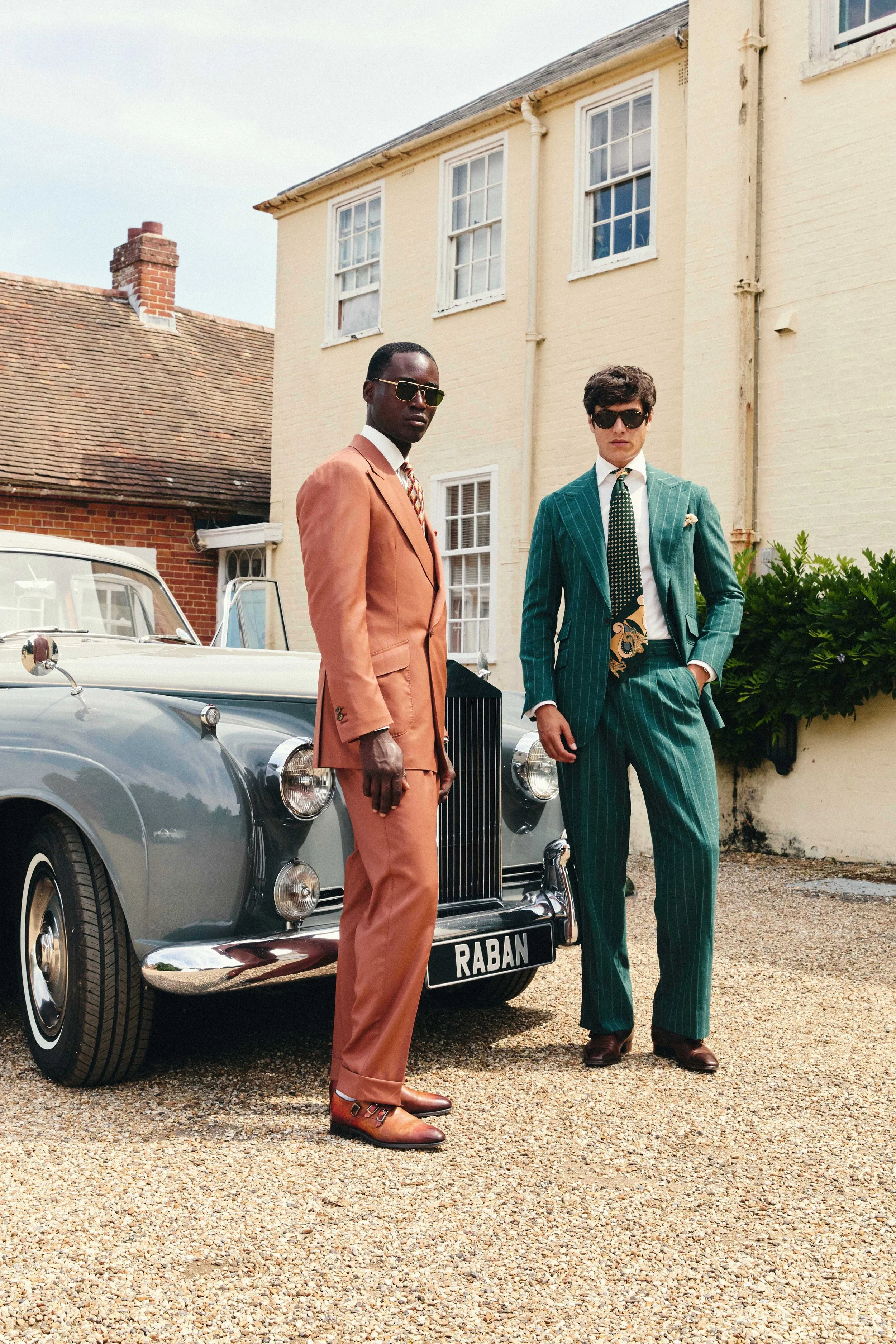 Two men dressed in vintage suits standing in front of a classic car outside a house with white walls and multiple windows. One man wears a peach-colored suit with matching trousers and brown shoes, while the other wears a green pinstripe suit with a white shirt and a patterned tie, both with sunglasses. The car has a visible license plate reading 'RABAN.'