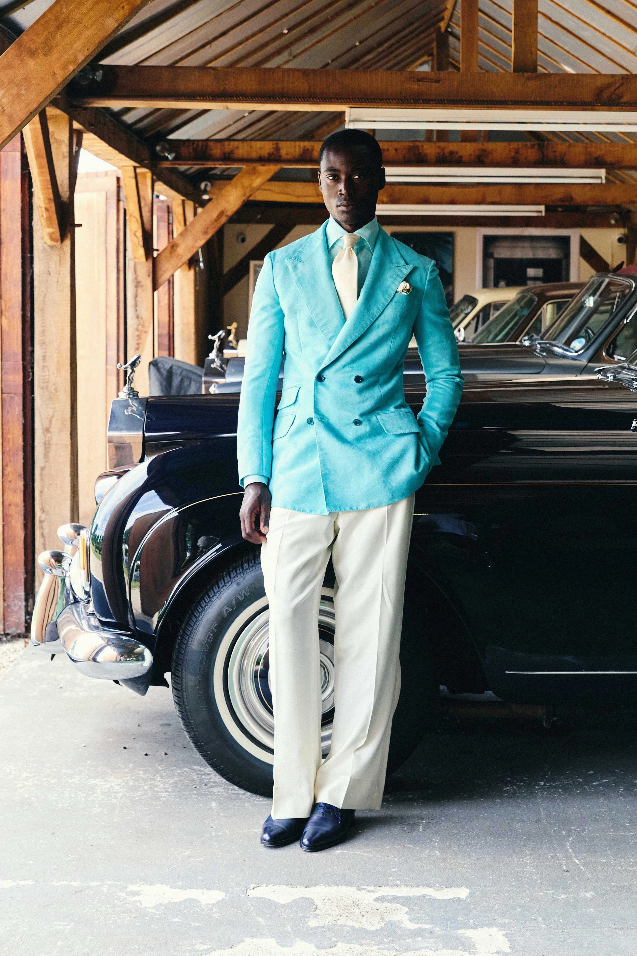 A man in a turquoise double-breasted blazer, cream trousers, and black shoes is standing in front of a black vintage car inside a wooden garage, with wooden beams and ceiling above.