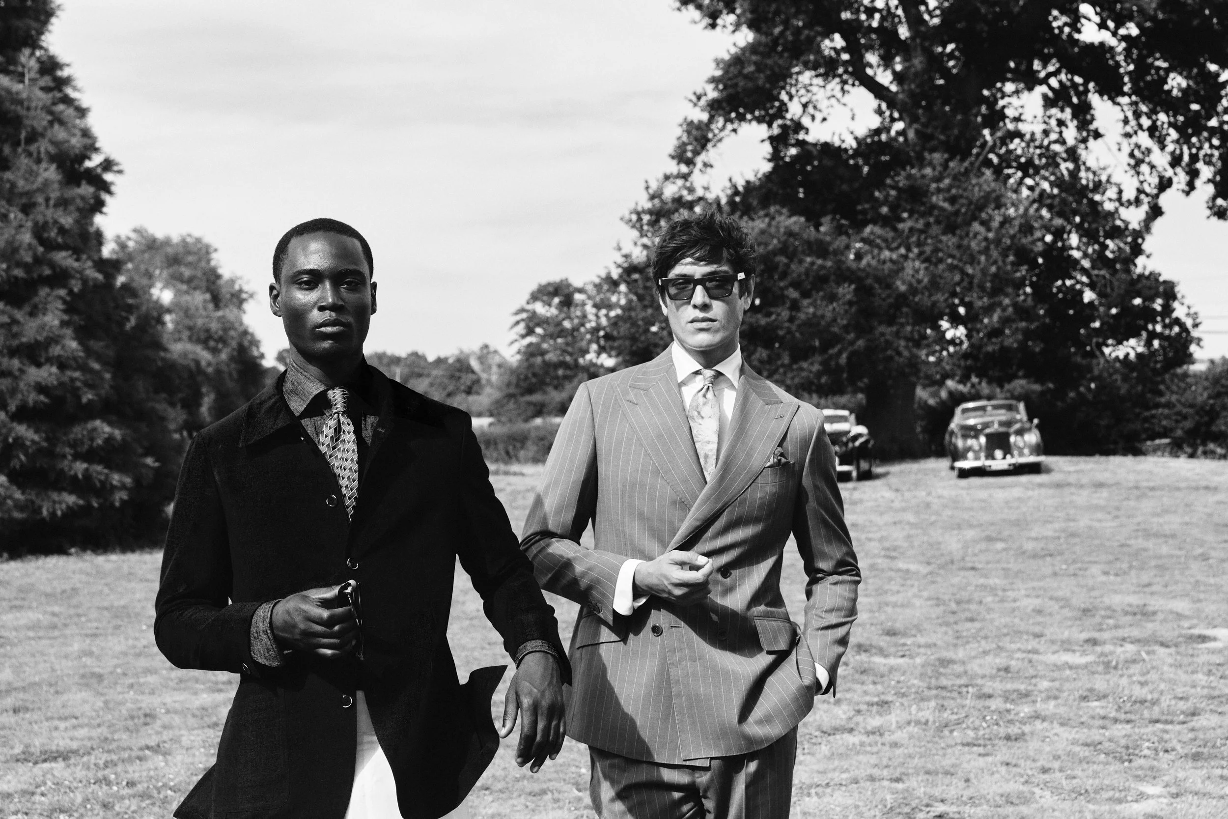 Two men walking outdoors, dressed in stylish suits, with a line of vintage cars in the background.