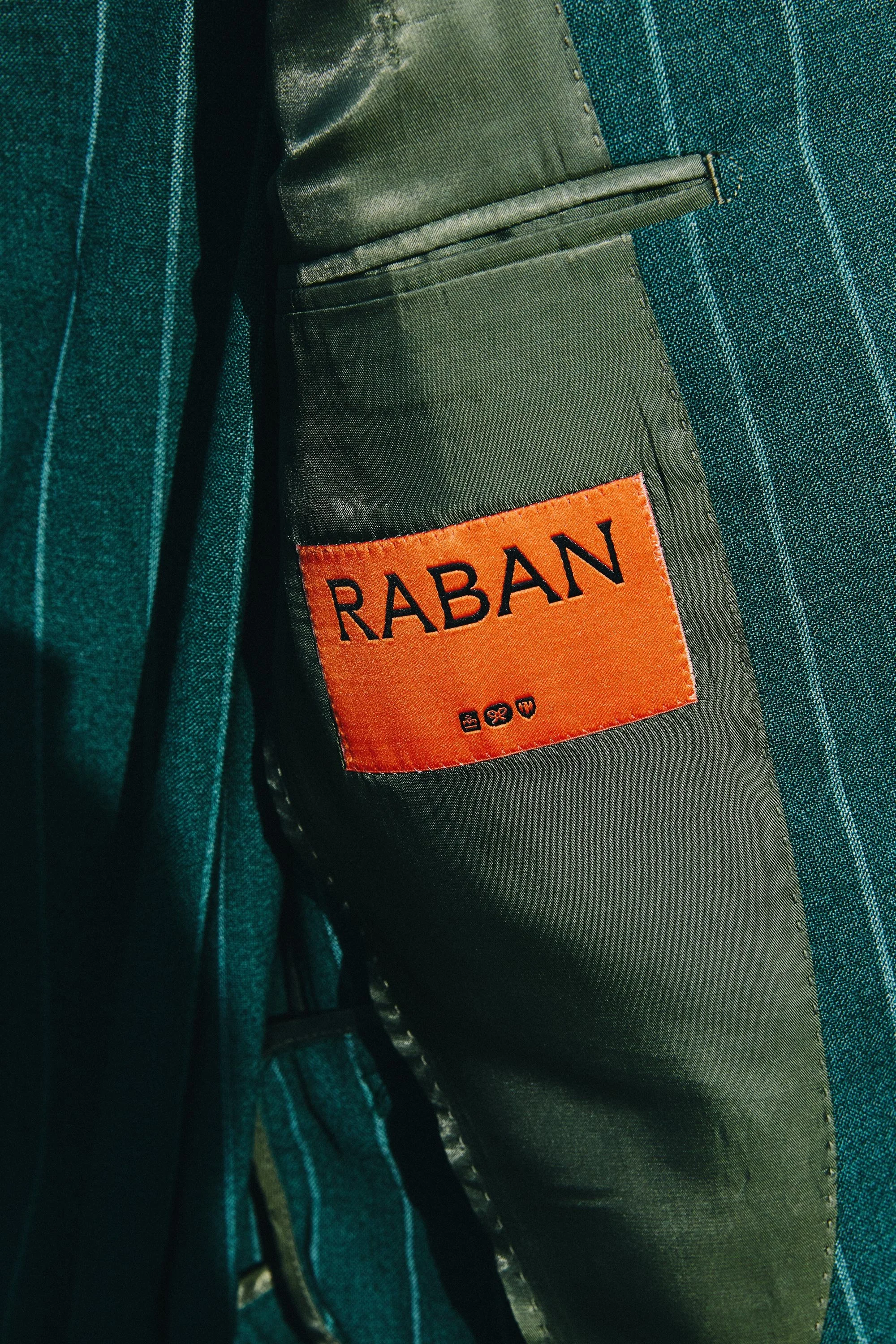 Close-up image of a suit jacket with a brown synthetic label that reads 'RABAN' in black letters, showing part of a person's arm and shirt with blue and green stripes.