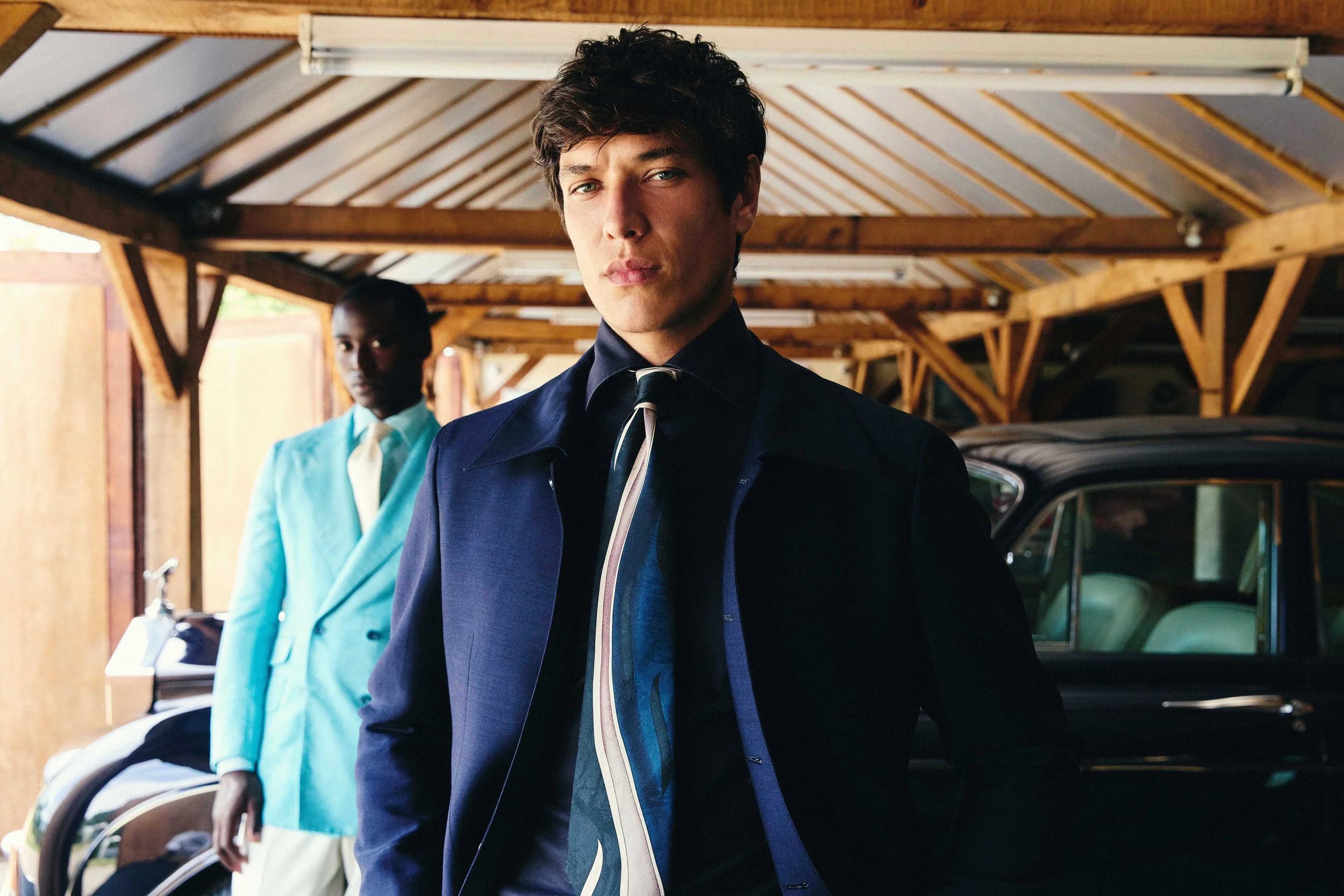 A young man with light skin and curly brown hair standing in front of a wooden garage, wearing a dark blue jacket with a reflective zipper and a dark shirt, with a serious expression. In the background, another person with dark skin, short hair, wearing a light blue suit, white shirt, and cream tie, stands next to a baby stroller.