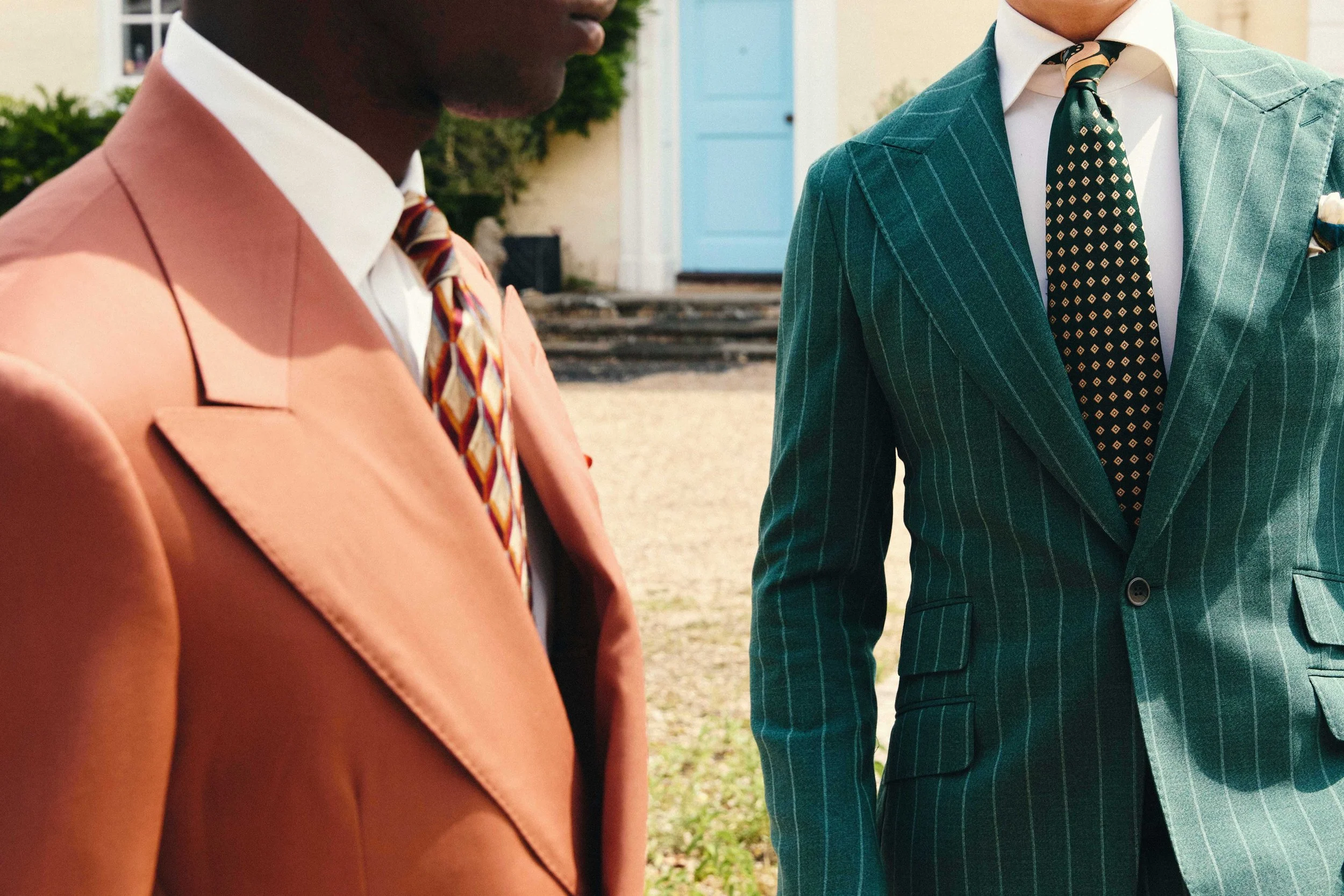 Close-up of two men dressed in colorful suits and patterned ties, standing outdoors near a building with a blue door and greenery.