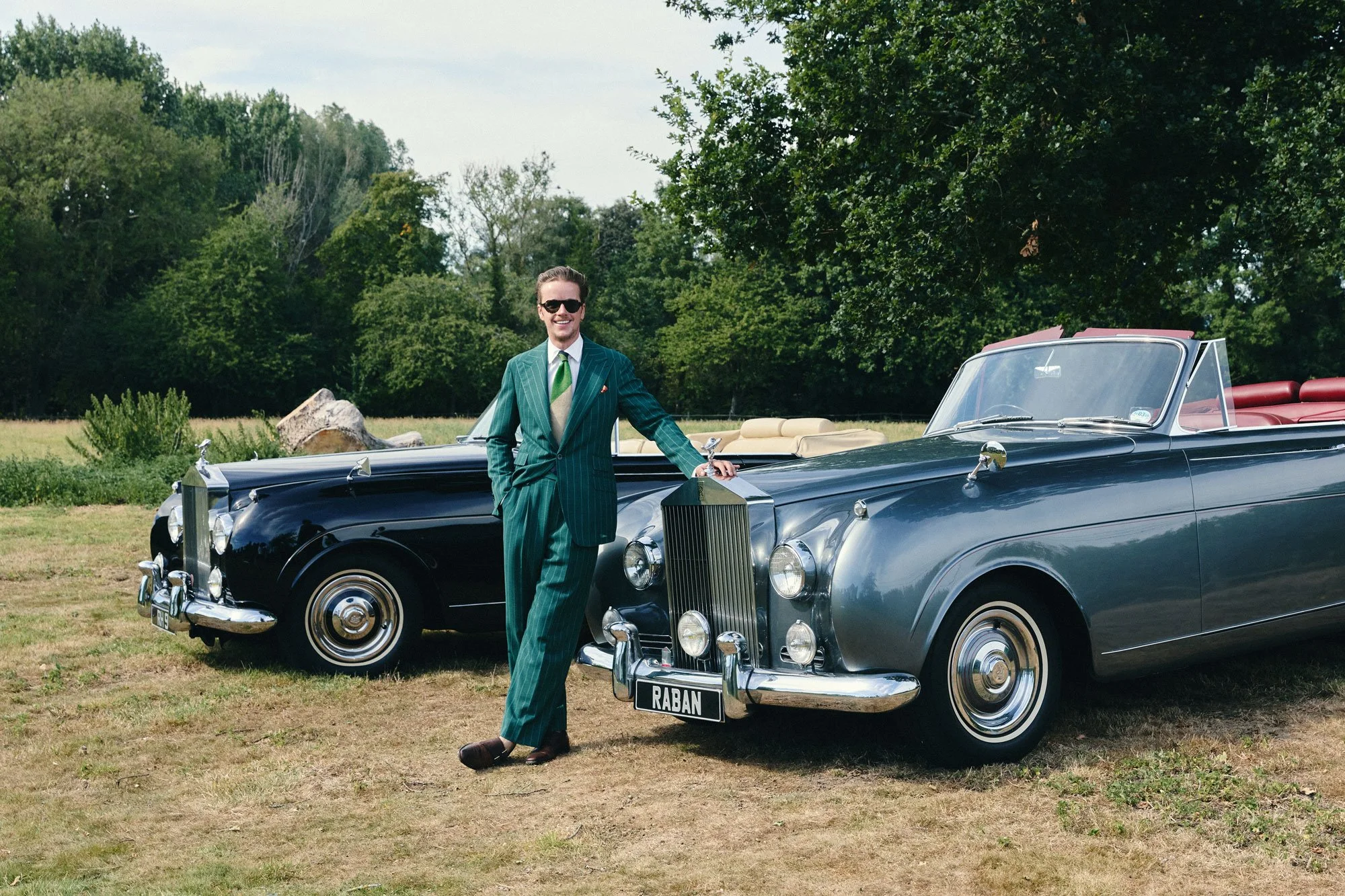 A man in a green striped suit and sunglasses standing next to a vintage black convertible car with a license plate that reads RABAN, parked on a grassy field with trees and a cloudy sky in the background.
