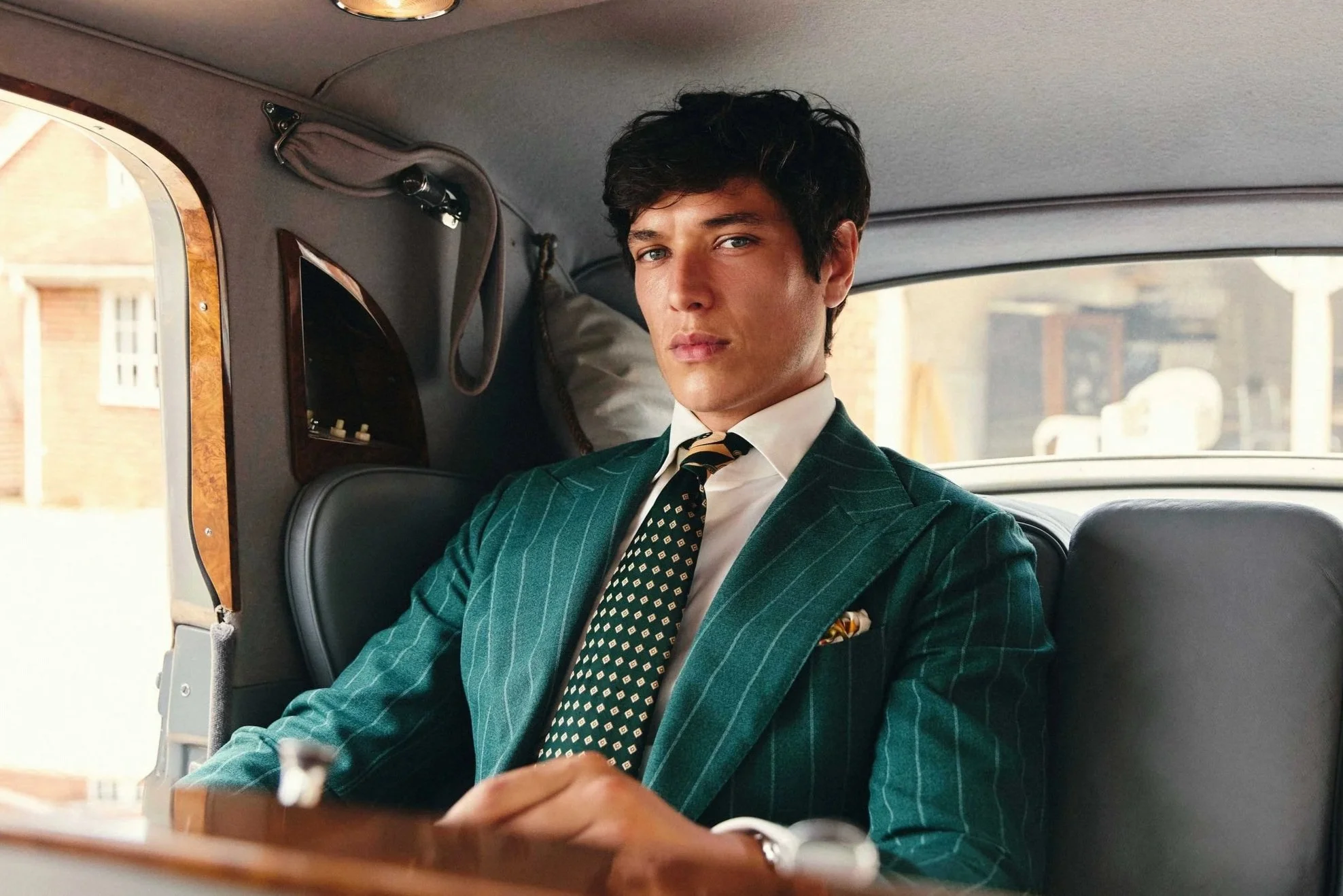 A young man with blue eyes and dark curly hair sitting in the back of a car, dressed in a green pinstripe suit, white shirt, and patterned tie, looking at the camera with a serious expression.