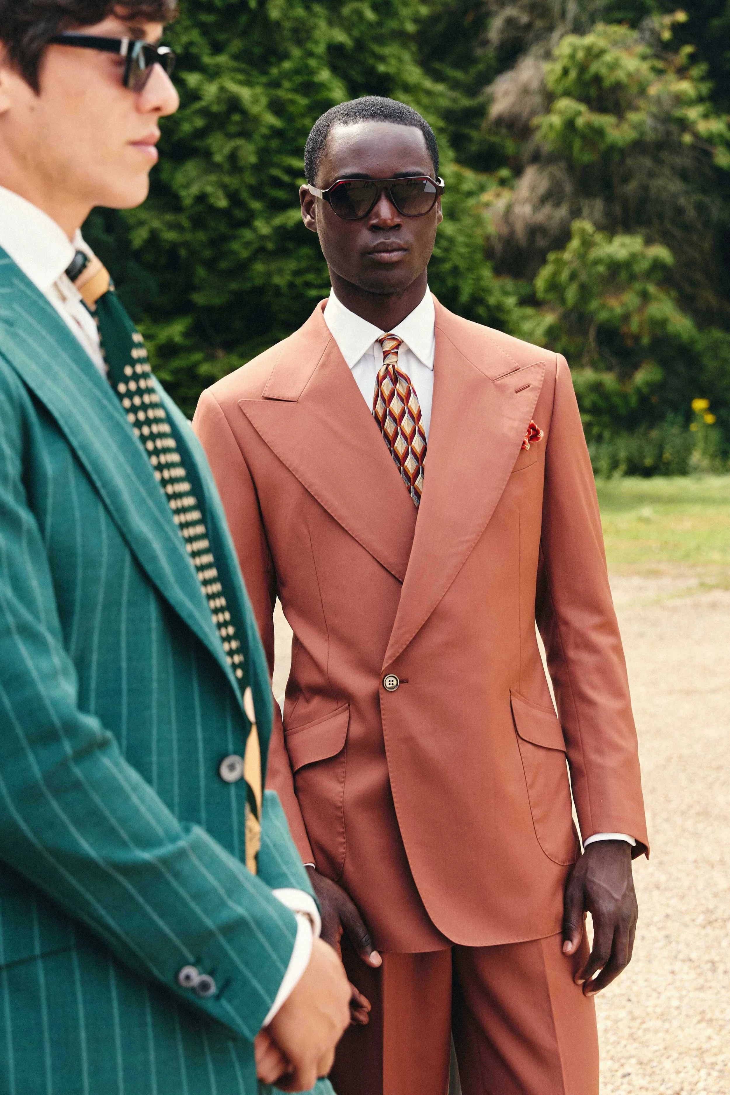 Two men wearing sunglasses and colorful suits stand outdoors with trees in the background. One man is in a teal pinstripe suit, and the other is in a rust-colored suit.
