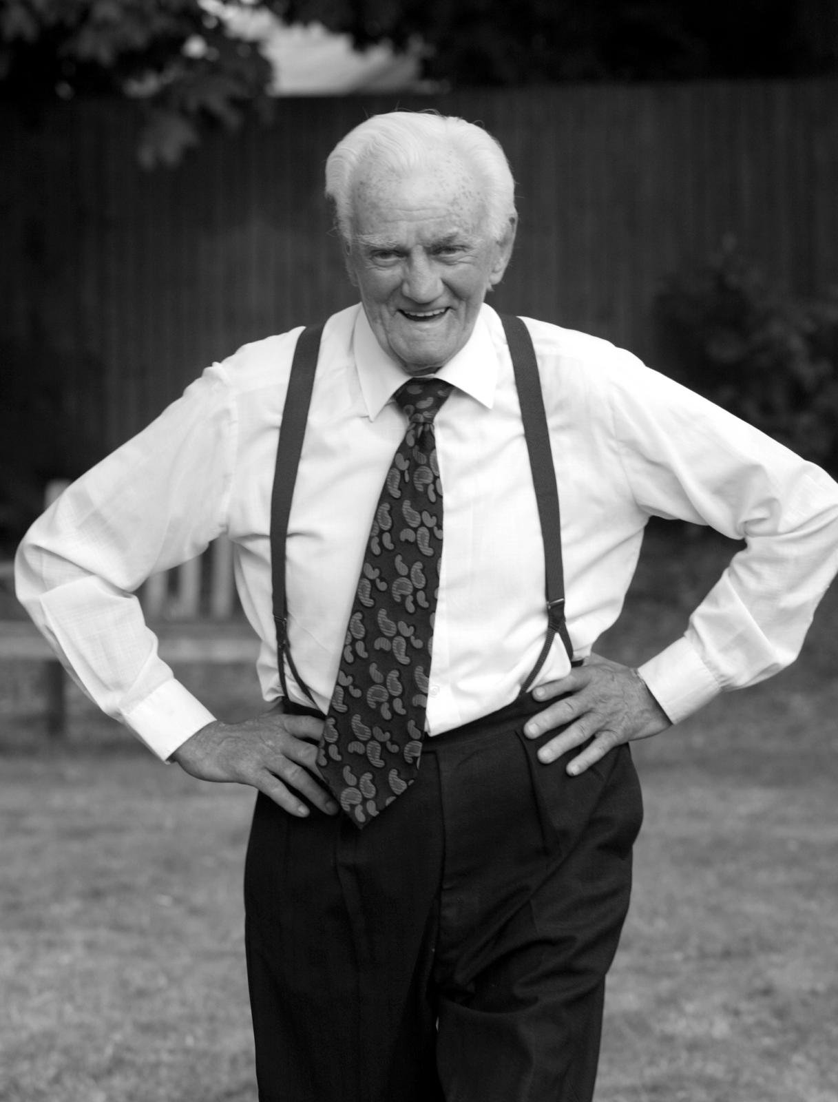 An elderly man in a white shirt, patterned tie, and suspenders stands outdoors with his hands on his hips, smiling and looking directly at the camera.