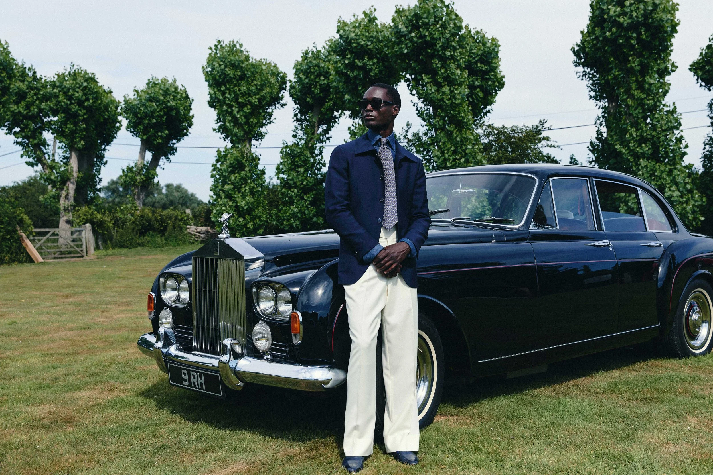 A man dressed in vintage fashion, wearing a dark blue jacket, patterned tie, white pants, and sunglasses, standing beside a classic black luxury car outdoors with a background of green trees and open sky.