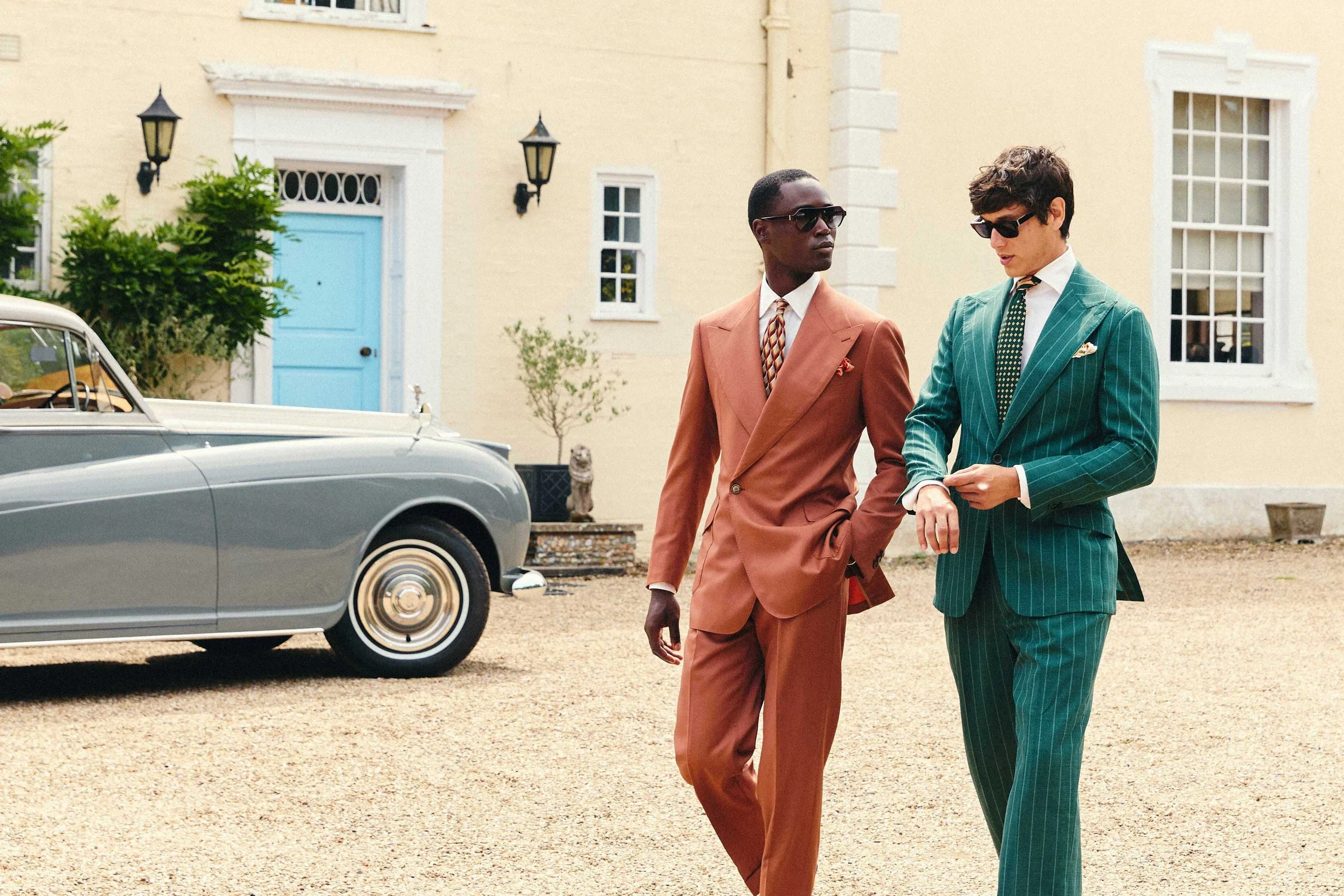 Two men in suits, wearing sunglasses, walking and talking outside a house with cream-colored walls, white window frames, and black outdoor lighting fixtures. A classic gray car is partially visible on the left.