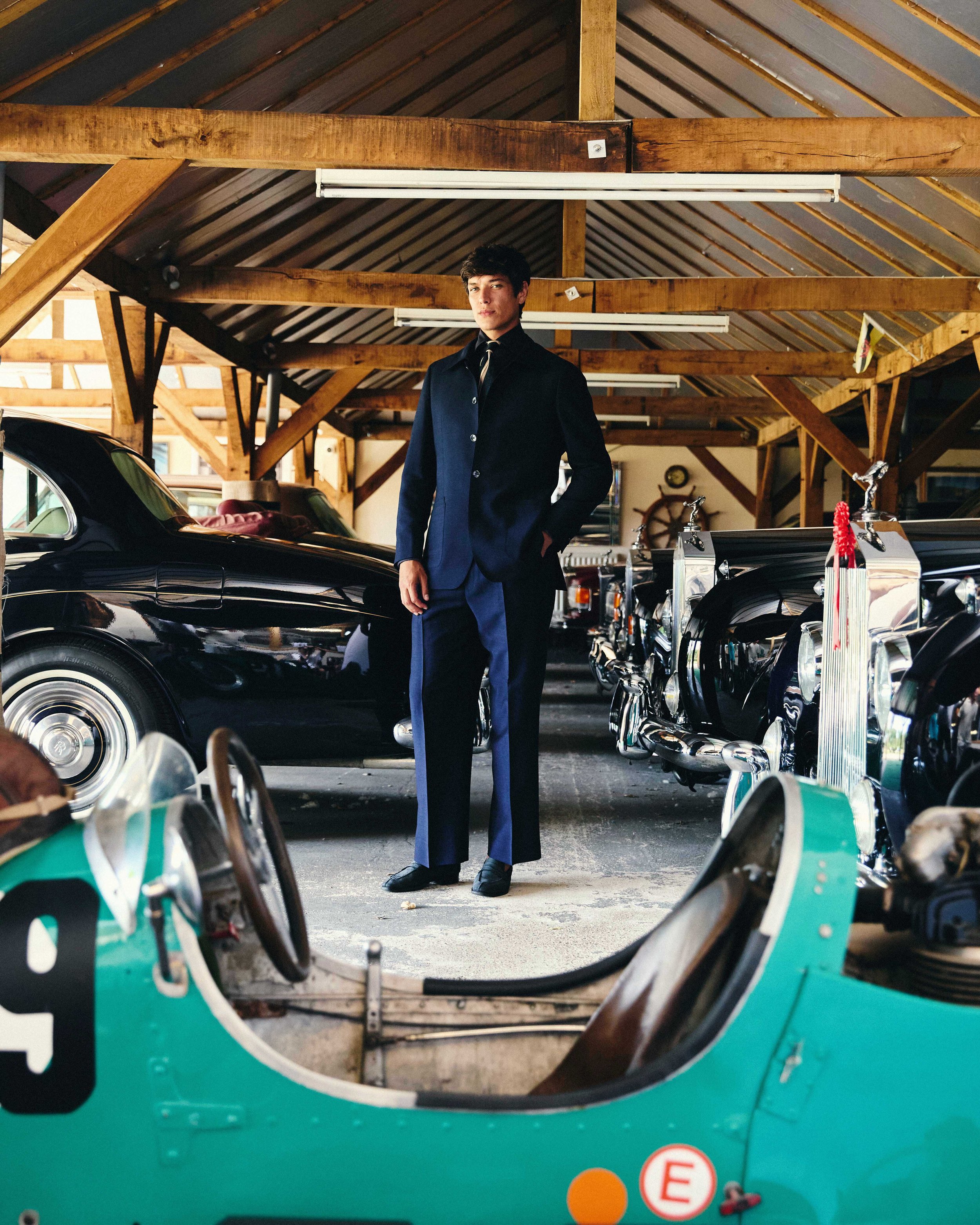 A young man in a dark suit standing inside a garage or warehouse, with several classic cars around him. The photo is taken through the windshield of a turquoise vintage car, which is in the foreground.